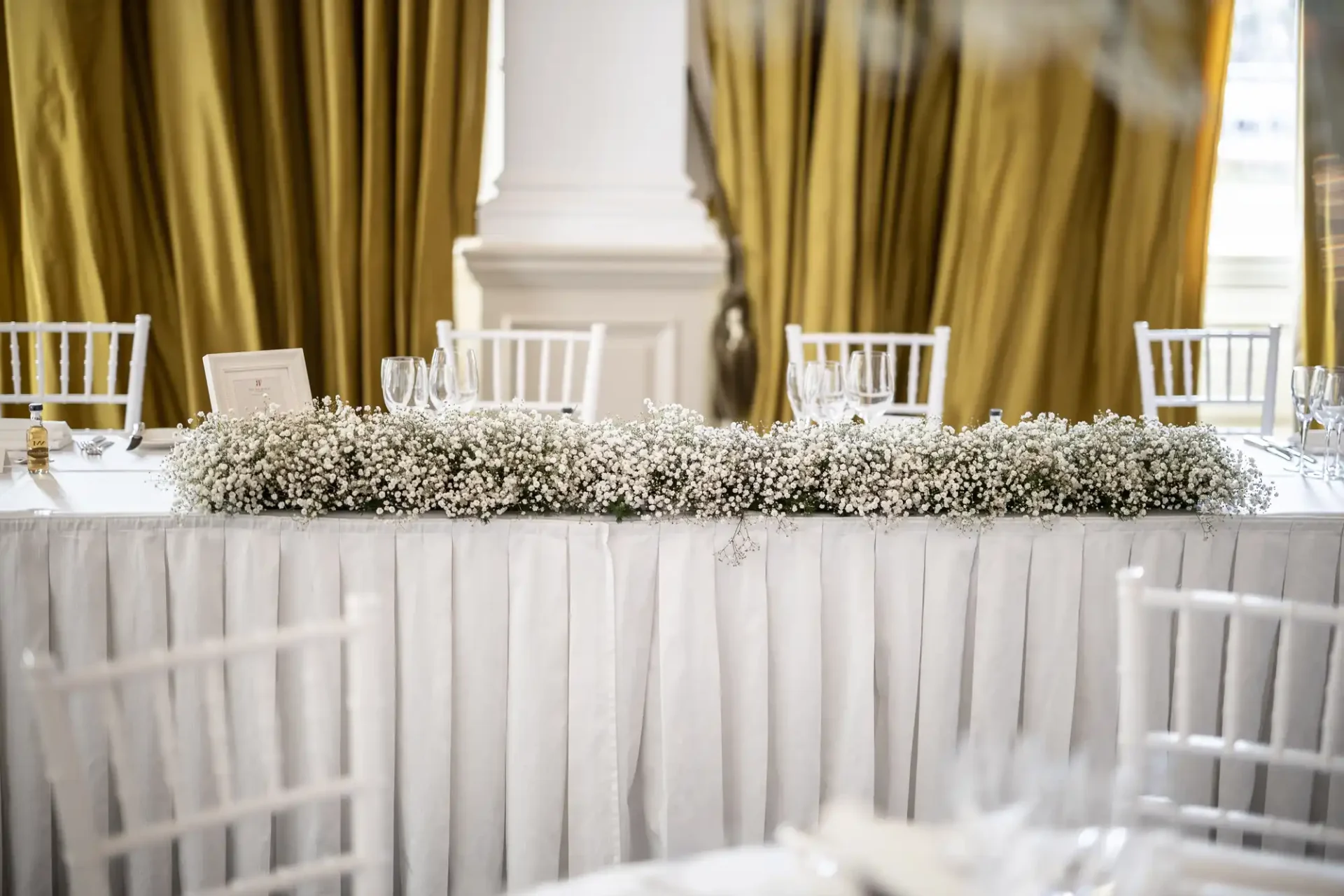 Elegant wedding table setting with white chairs, long floral centerpiece, and golden curtains in a luxurious room.