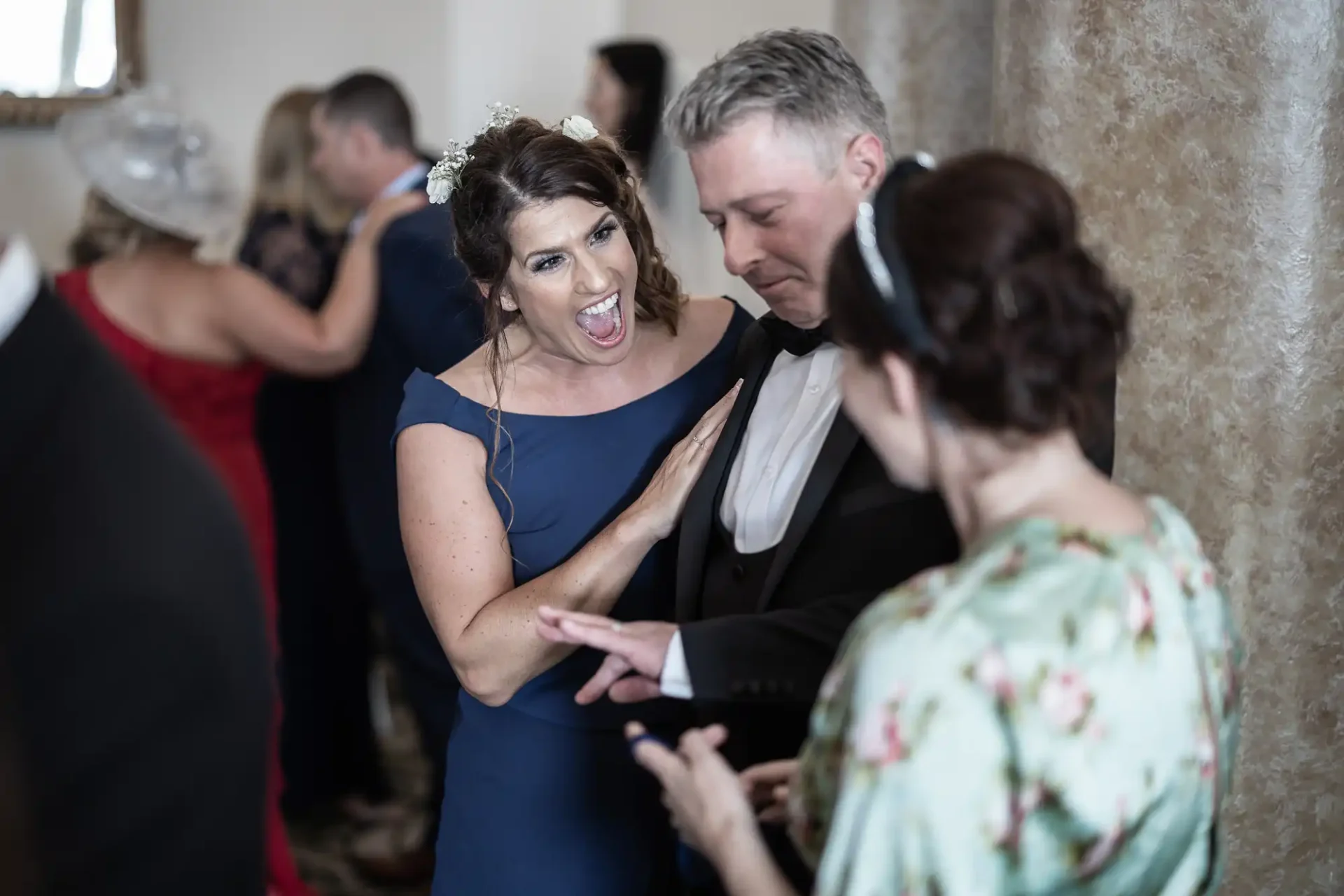 Woman in a blue dress playfully shows her muscle to a smiling man in a tuxedo, while another woman in a floral dress watches at a social event.