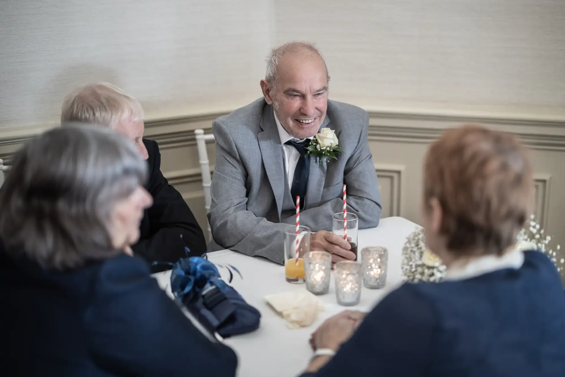 Elderly man with a boutonniere laughing at a table with three other guests at a formal event, featuring candles and drinks with straws.