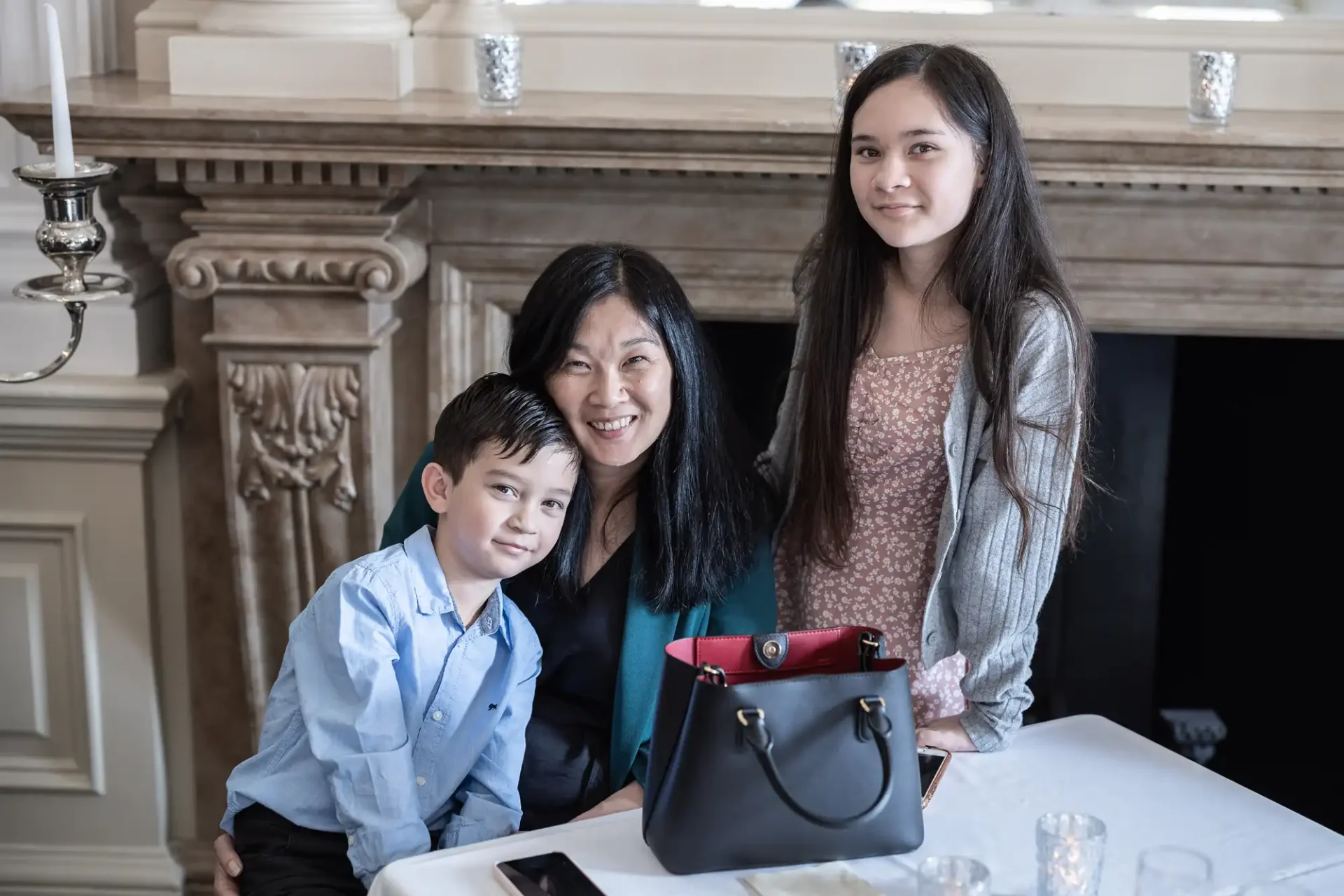 A woman and two children smiling at a dining table in an elegant room, with a stylish handbag placed on the table.