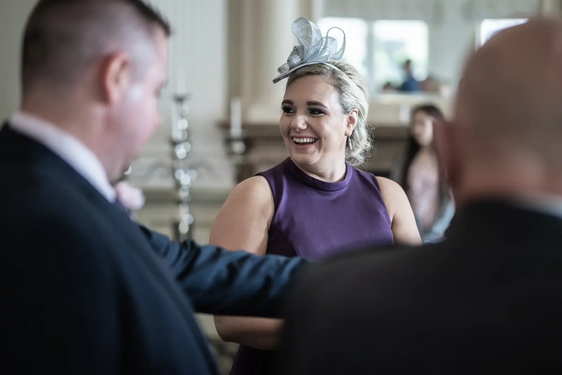 A woman in a purple dress and gray fascinator smiles while talking to two men in a bright, elegant room.