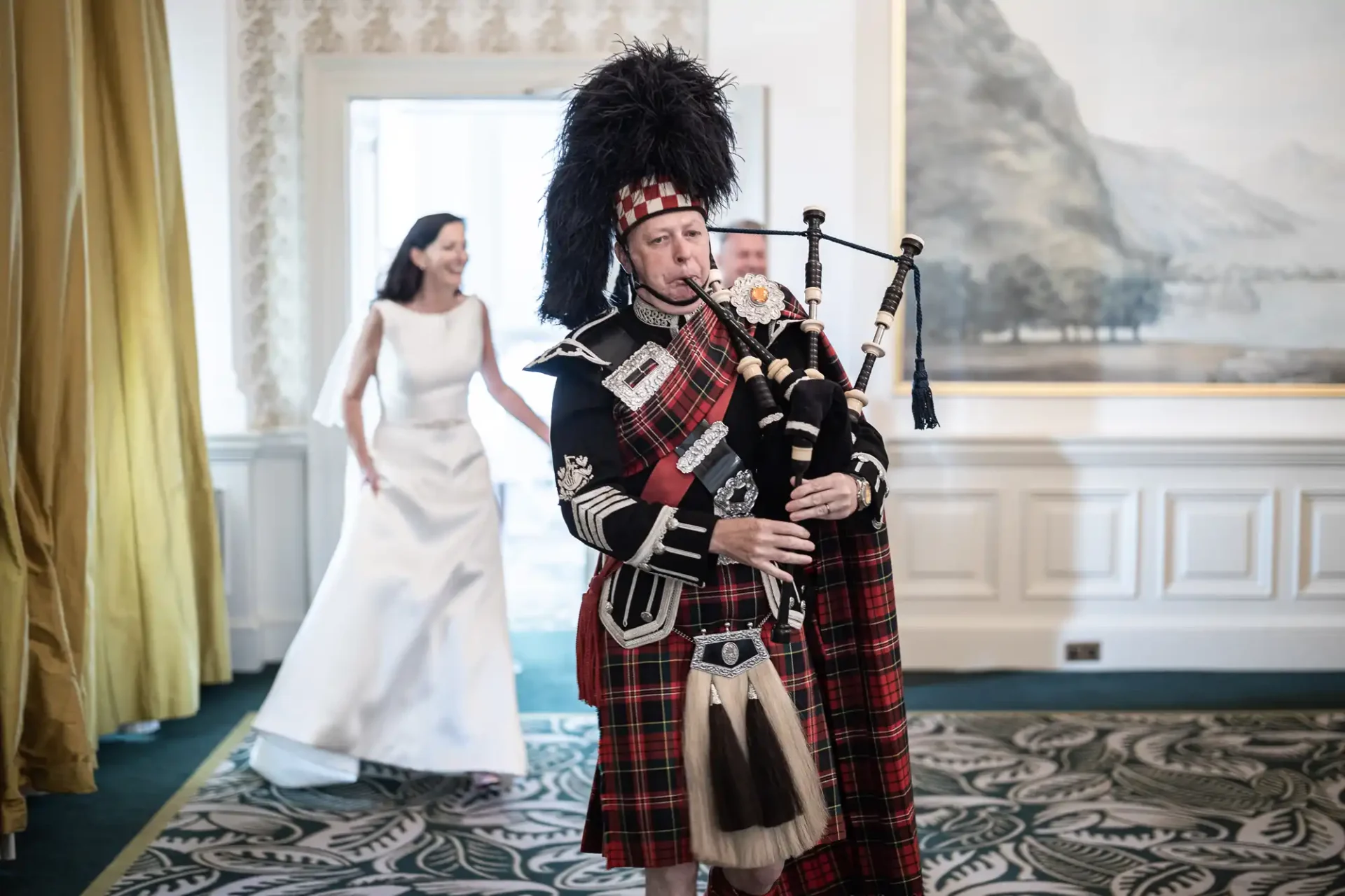 A bagpiper in traditional scottish attire plays while a bride in a white dress smiles in the background in an elegantly decorated room.