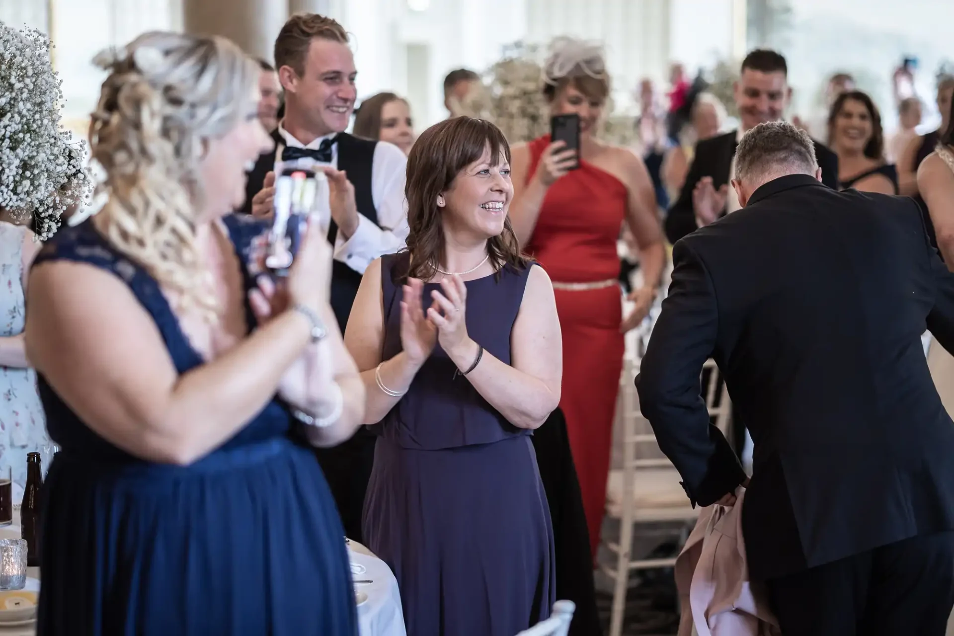 Guests smiling and clapping at a wedding reception, with some capturing the moment on their phones.