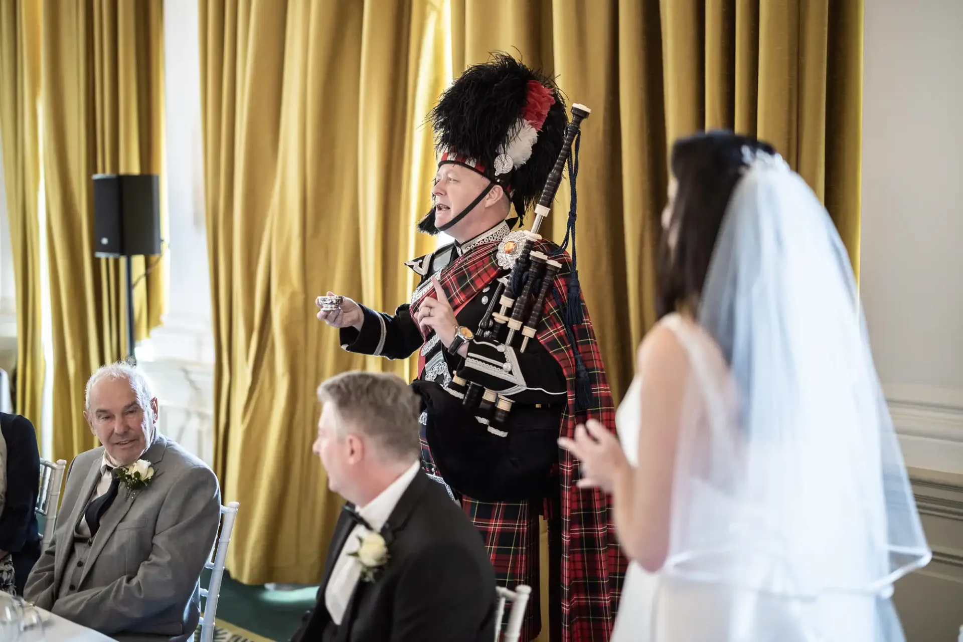 A bagpiper in traditional scottish attire, including a tartan kilt, addresses a bride and guests at a wedding reception indoors.