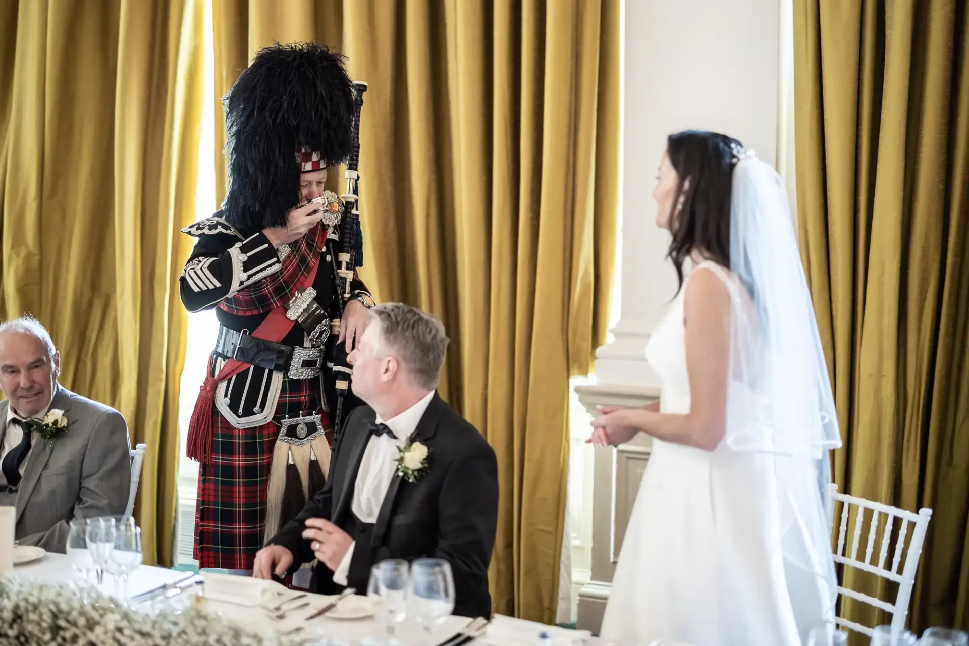 A bagpiper in traditional scottish attire plays at a wedding reception, entertaining a smiling bride and groom seated at a table.
