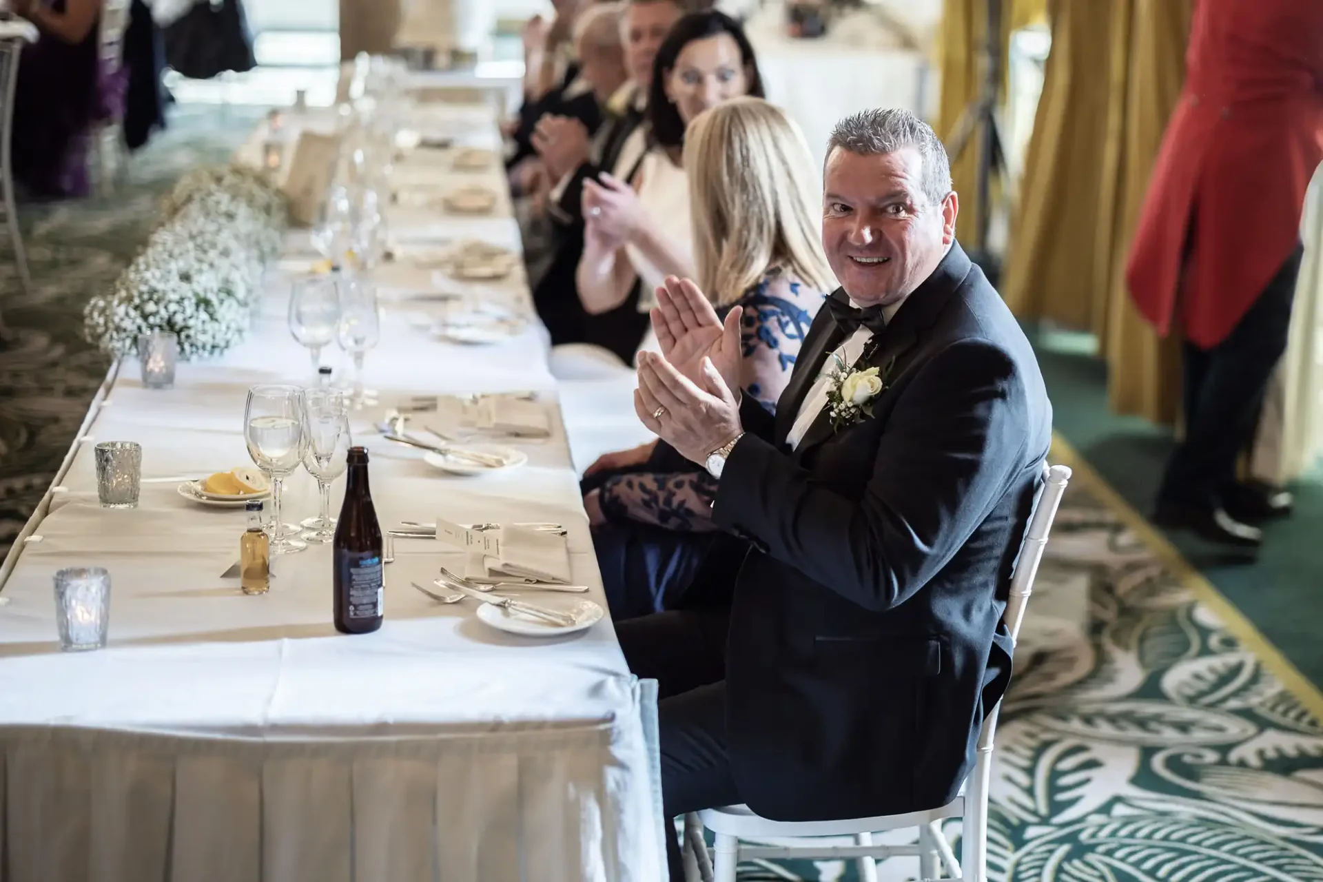 A man in a suit claps at a formal dining table during an event, surrounded by other guests.