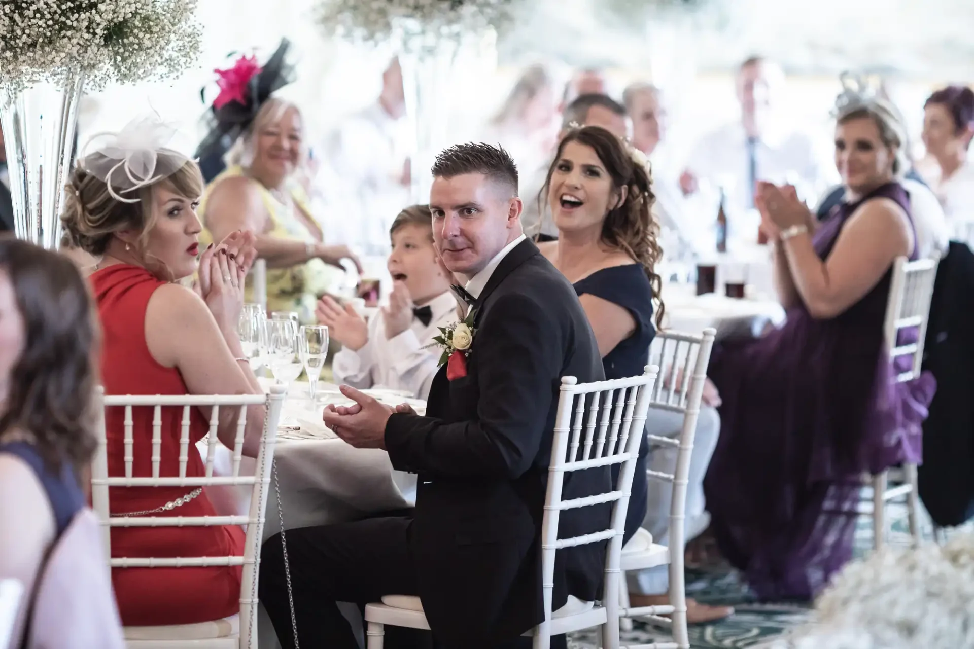 A wedding reception scene with guests in formal attire, focusing on a man in a suit and woman in a fascinator, both seated and clapping.