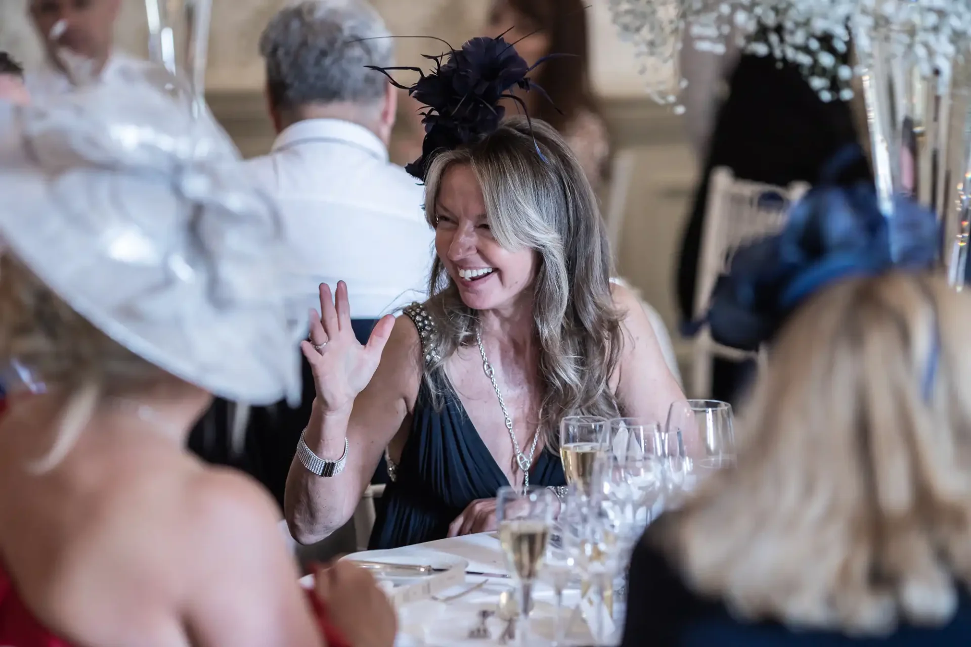 A woman, wearing a floral dress and a black fascinator, laughs and waves at someone across a table during a formal event with other guests in hats.