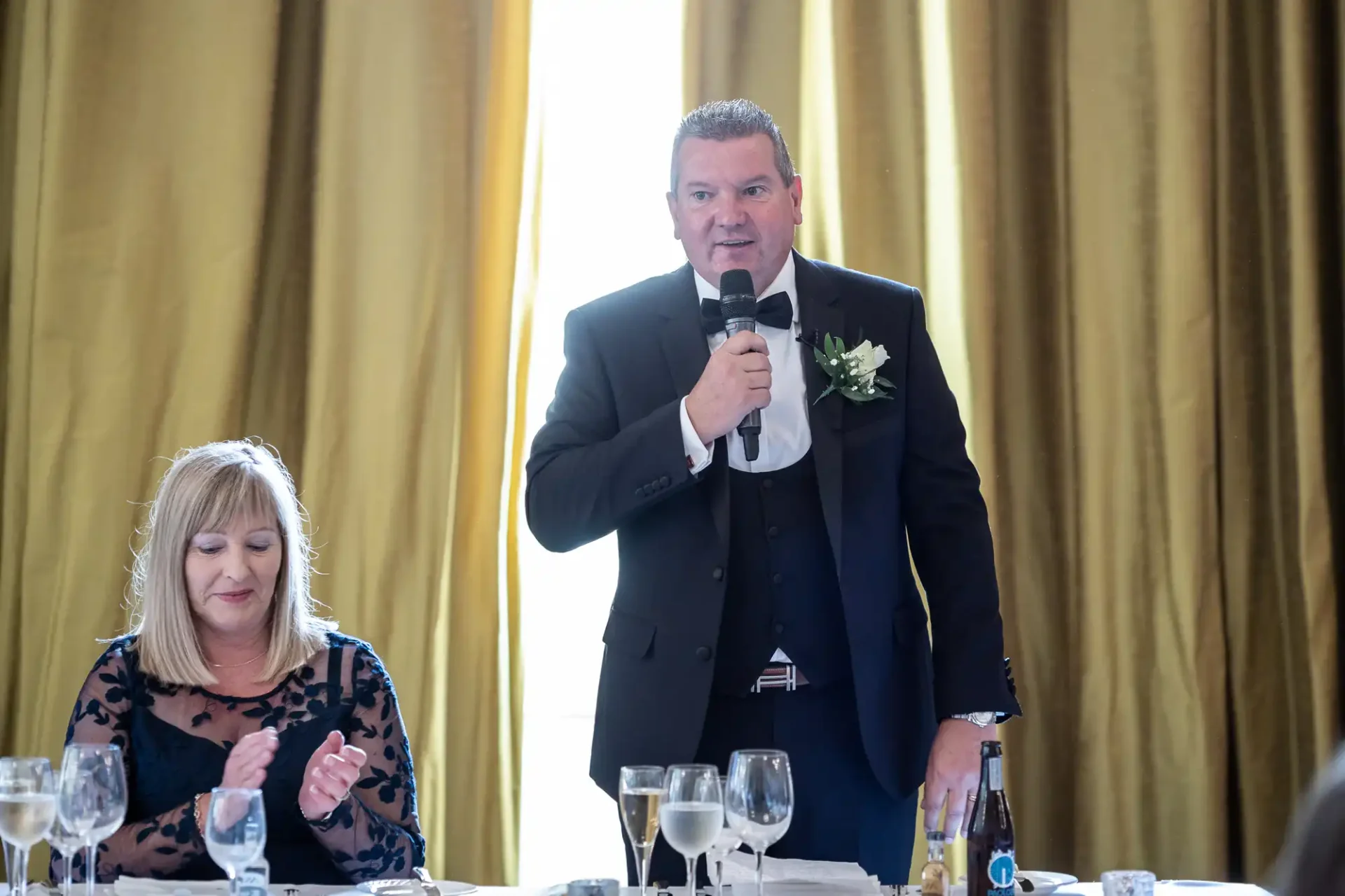 Man in formal attire delivering a speech with a microphone at a wedding reception, with a seated woman nearby.