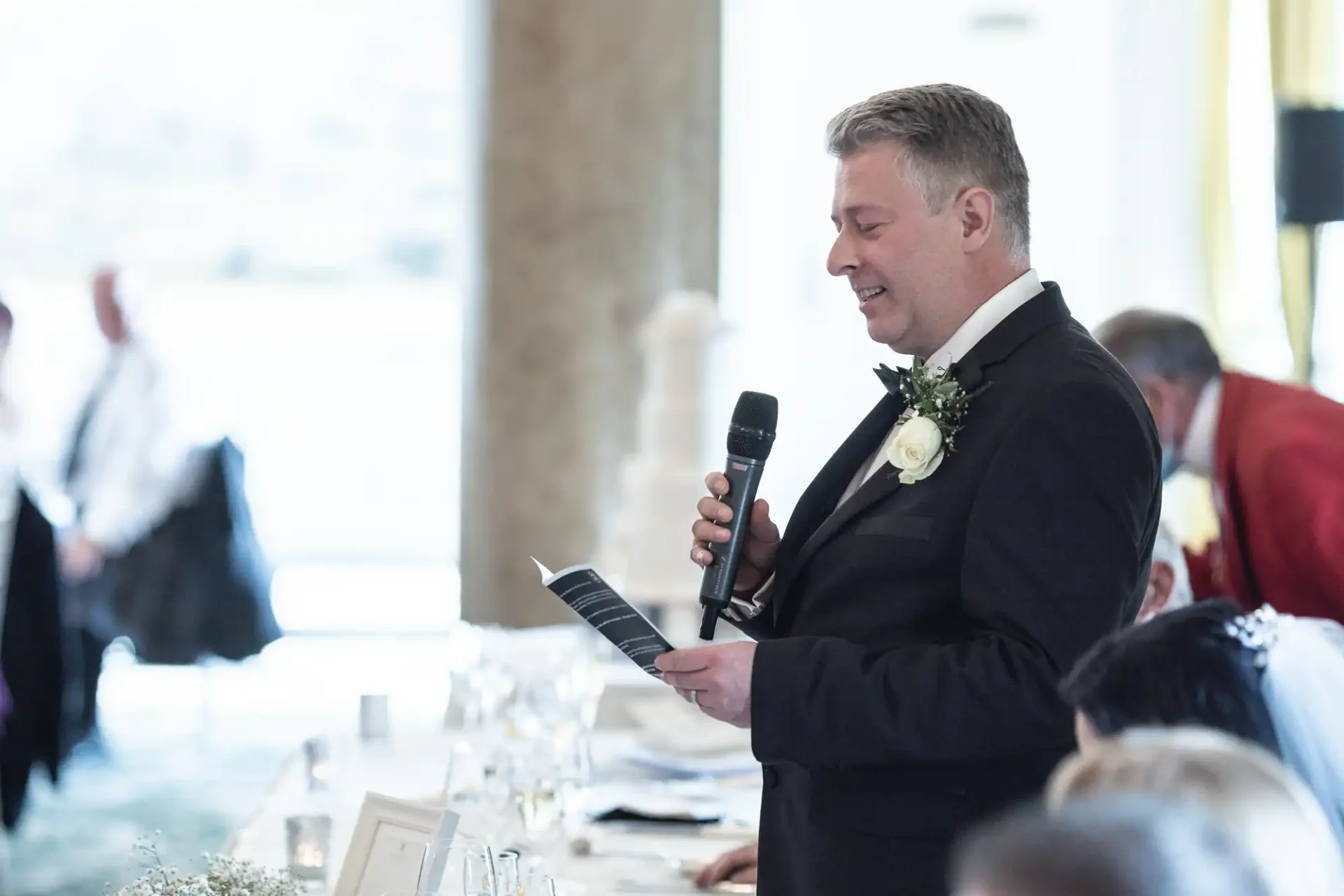 A man in a wedding suit gives a speech holding a microphone and a sheet of paper at a banquet hall.