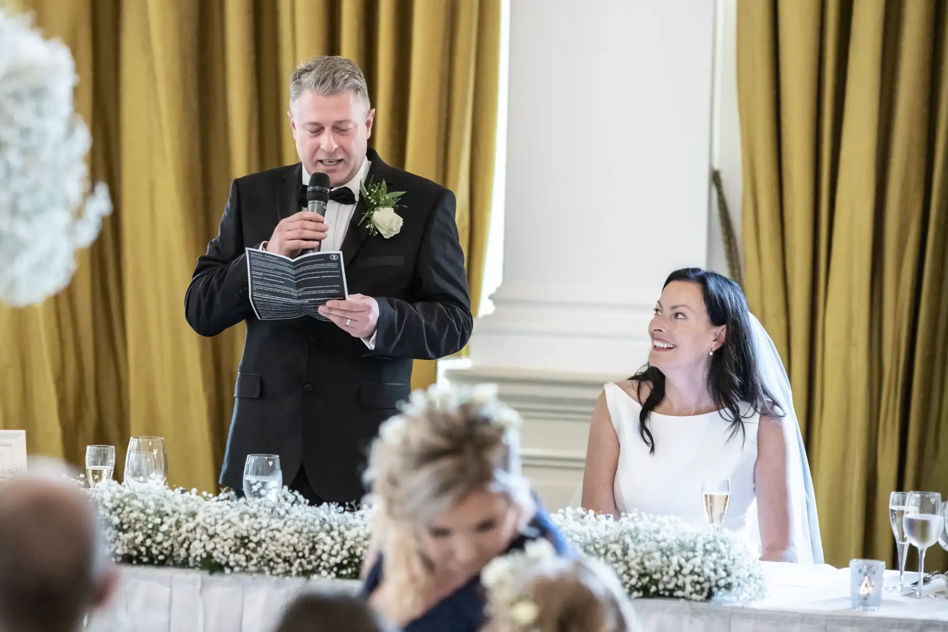 A man in a suit giving a speech at a wedding reception, holding a notebook, as a smiling bride in a white dress listens, surrounded by guests and floral decorations.