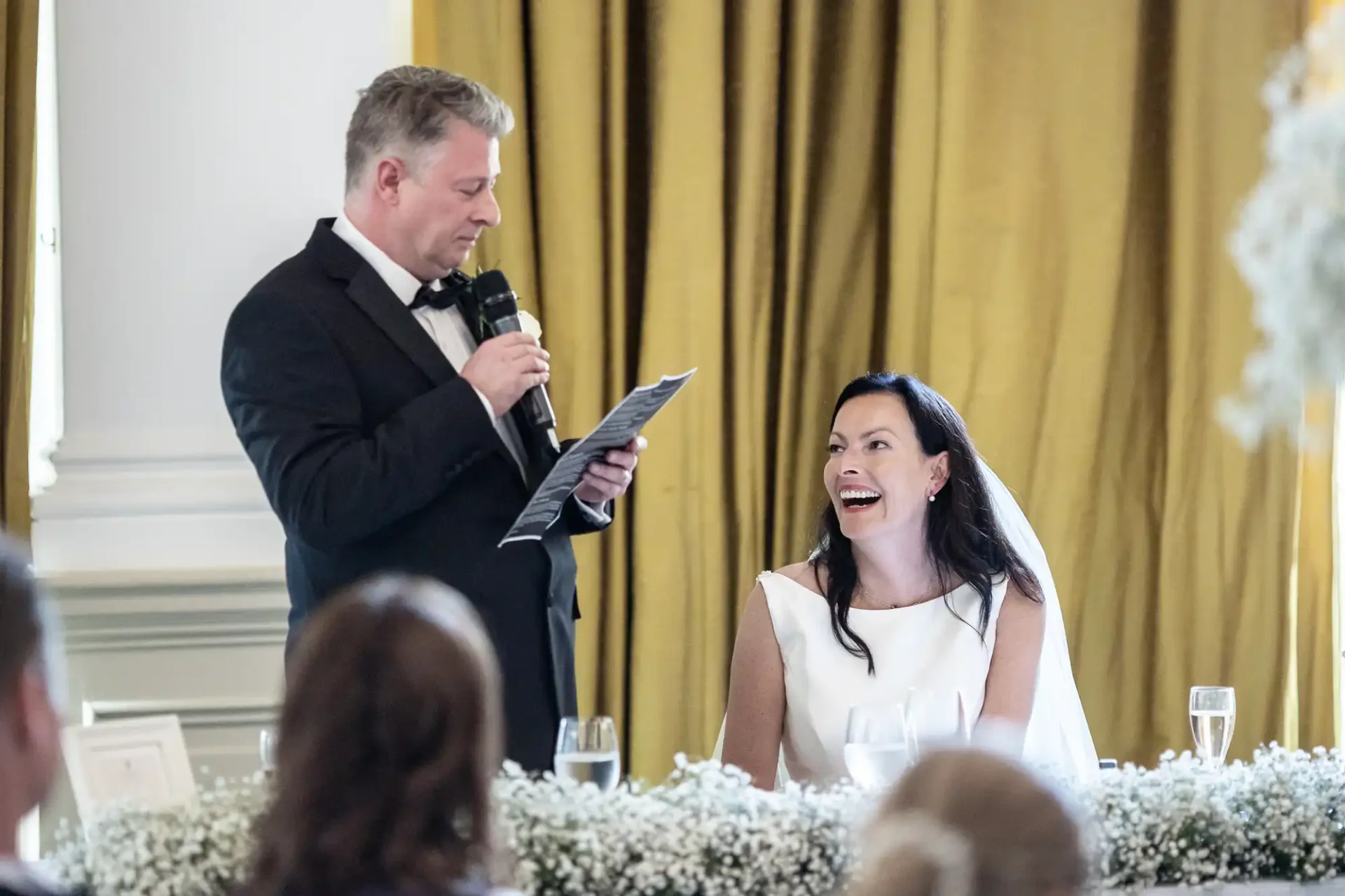 A bride laughs joyfully during a speech given by a man in a tuxedo at a wedding reception, with guests and floral decorations visible.