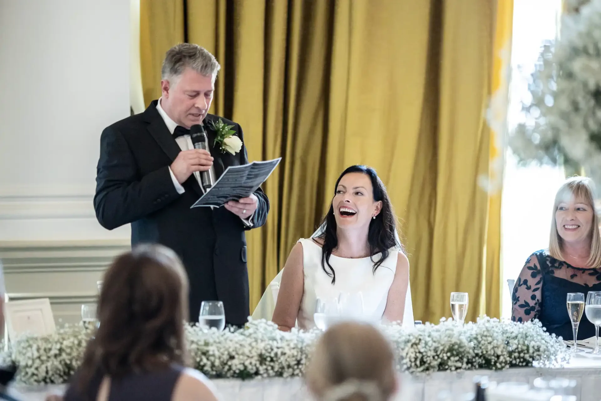 A man in a suit gives a speech holding papers at a wedding, eliciting laughter from the bride and other female guest seated at a decorated table.