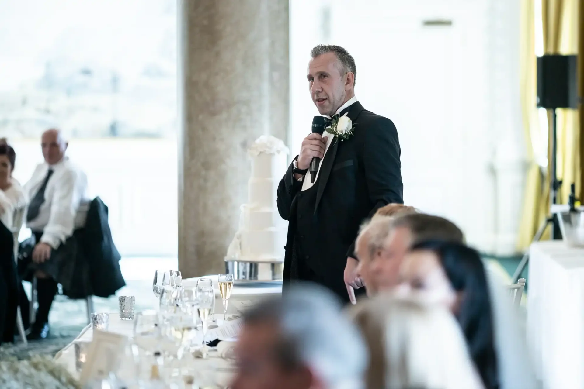 Man in suit giving a speech at a wedding reception, holding a microphone, with guests seated at tables.