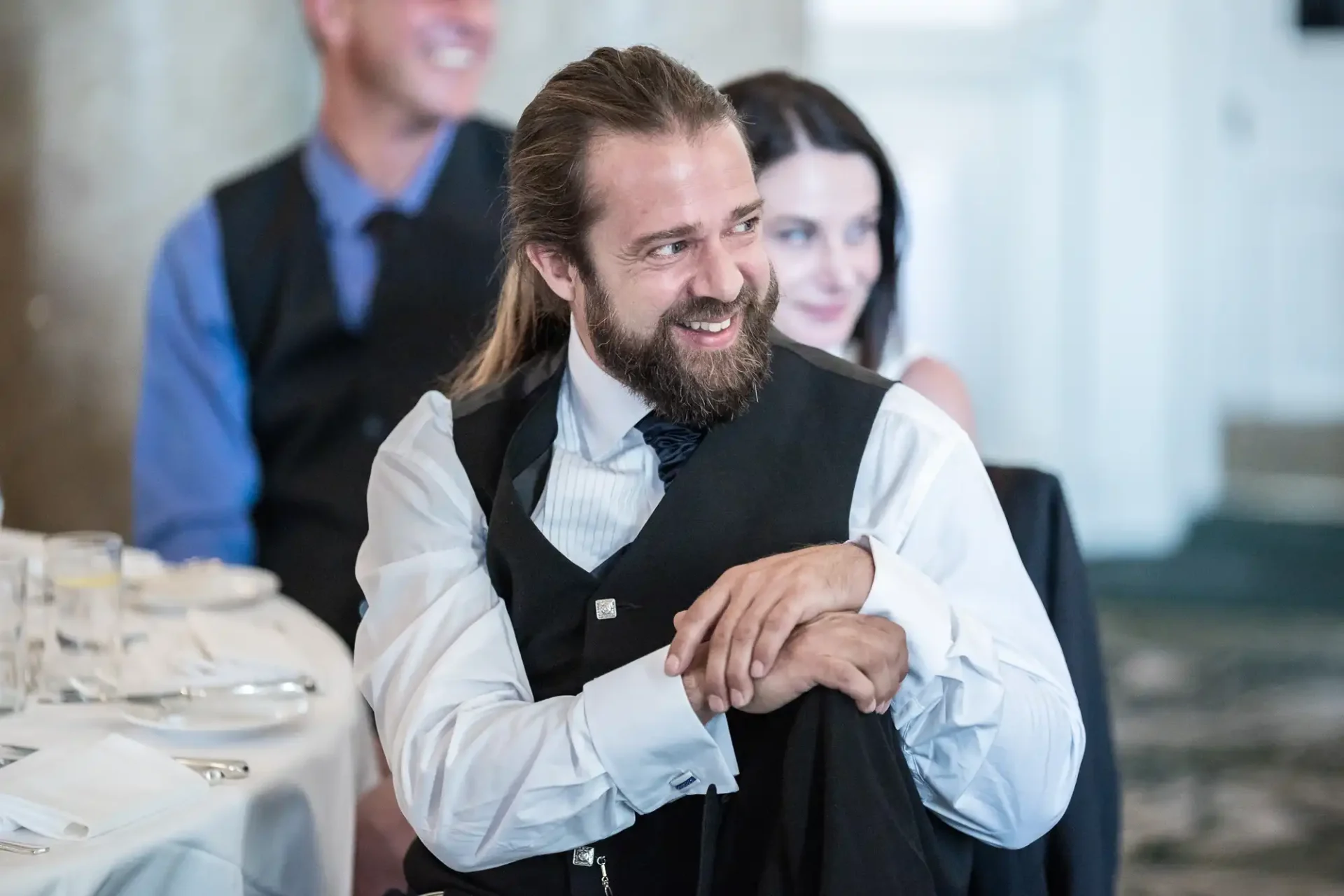 A man with a beard, wearing a vest and tie, smiles warmly at a wedding reception, with guests visible in the background.