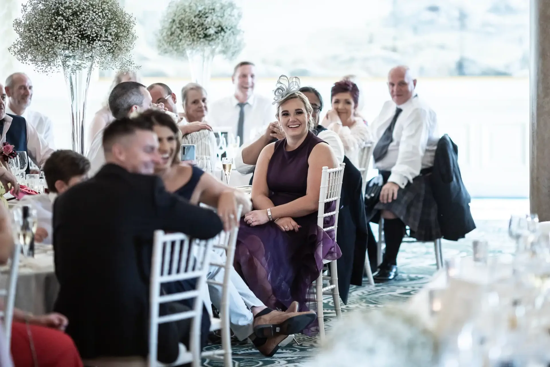 Woman in purple dress smiling at a wedding reception, seated at a table surrounded by guests.