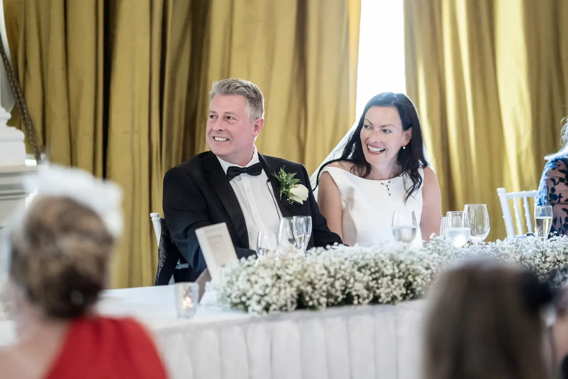 A bride and groom smiling joyfully at a wedding reception table, adorned with white flowers.