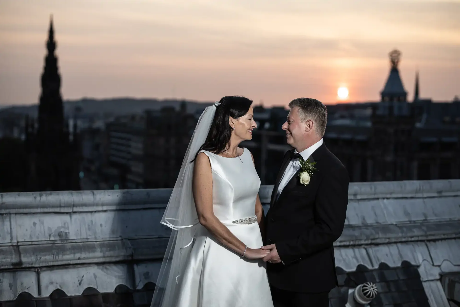 Bride and groom hold hands on a rooftop at sunset, with city skyline including a historic tower in the background.