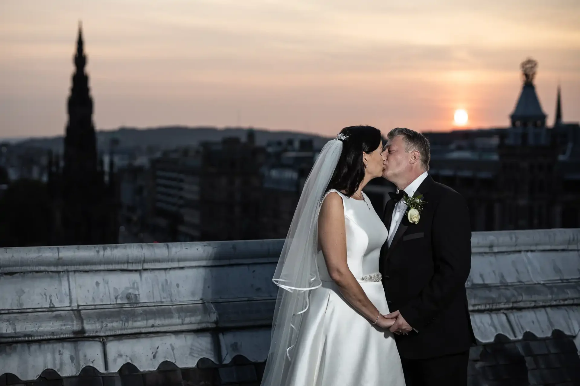 A bride and groom kissing at sunset on a rooftop with city skyline views.