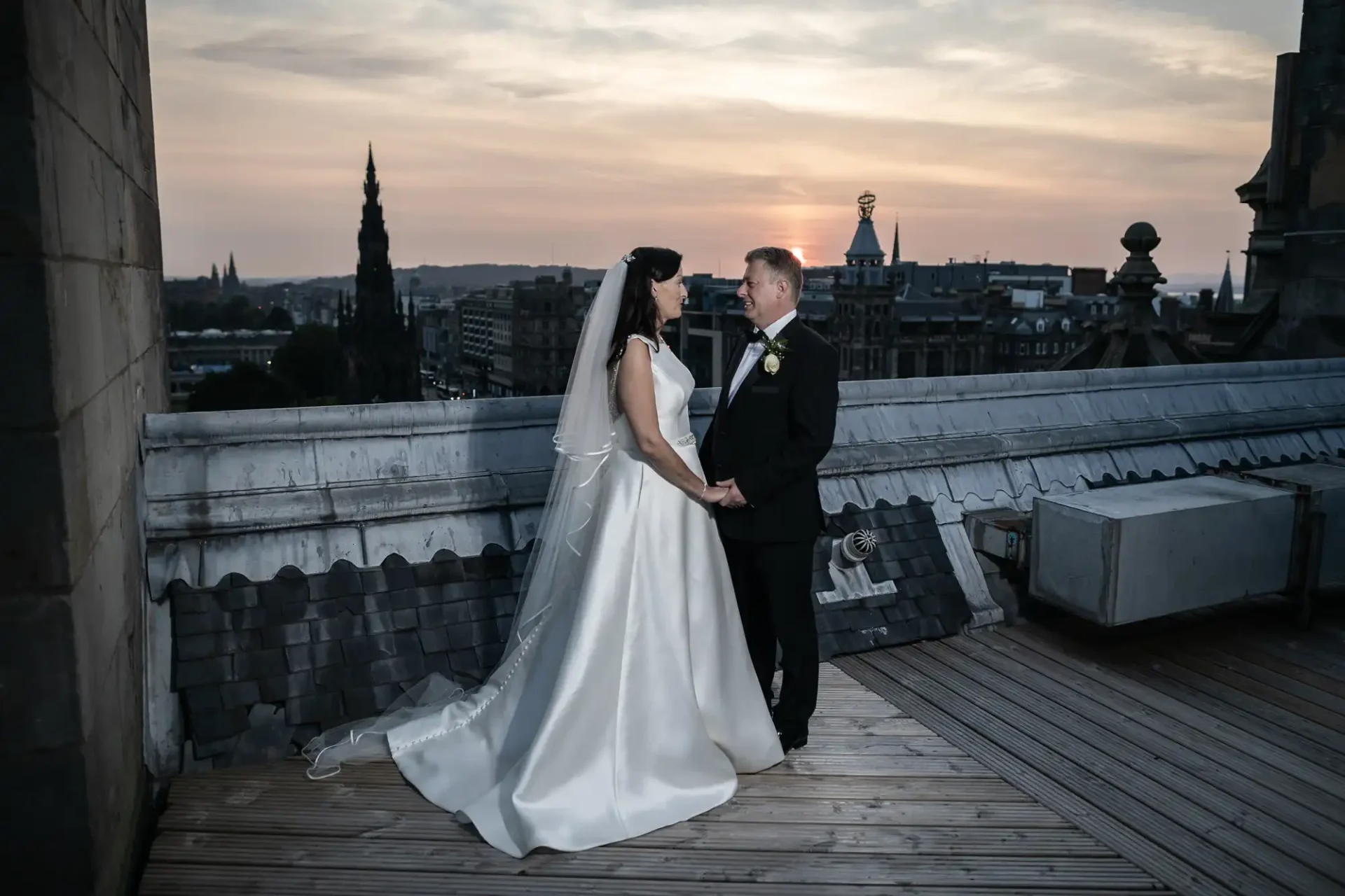 A bride and groom holding hands on a rooftop at dusk, with a cityscape in the background.