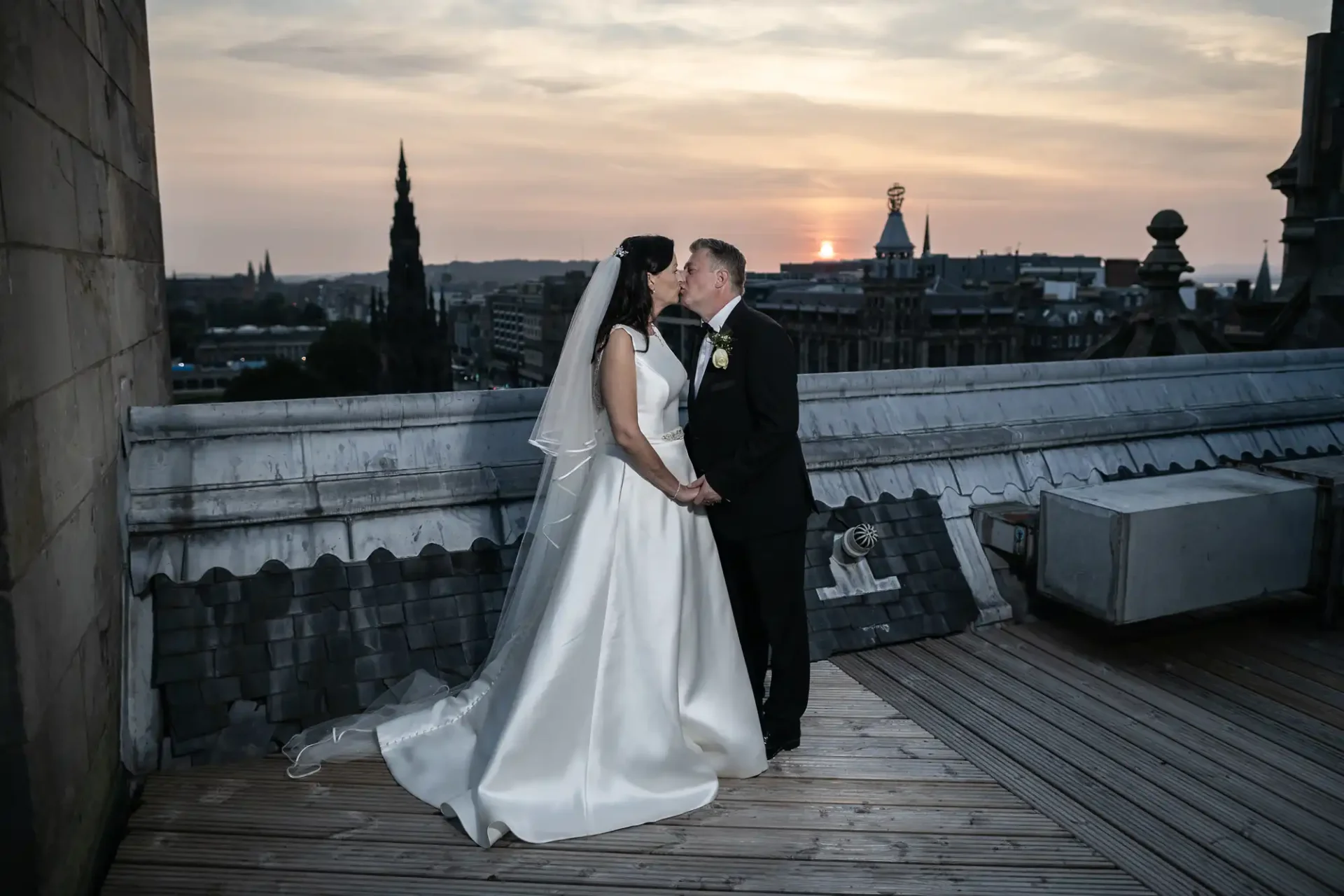 Bride and groom holding hands and kissing at sunset on a city rooftop with skyline in the background.