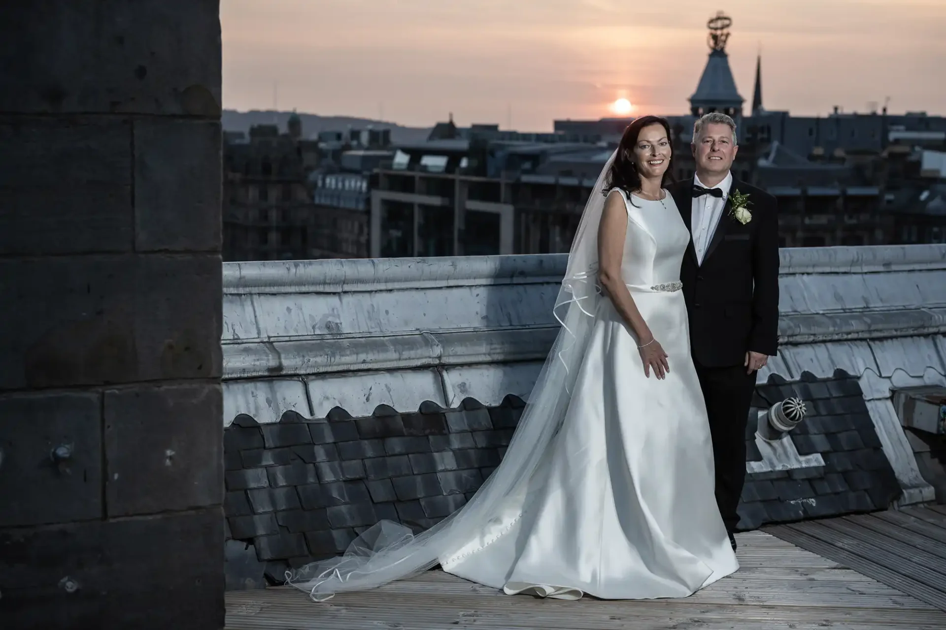 A bride and groom stand on a rooftop at sunset, with city buildings in the background. the bride wears a white dress and the groom a dark suit.