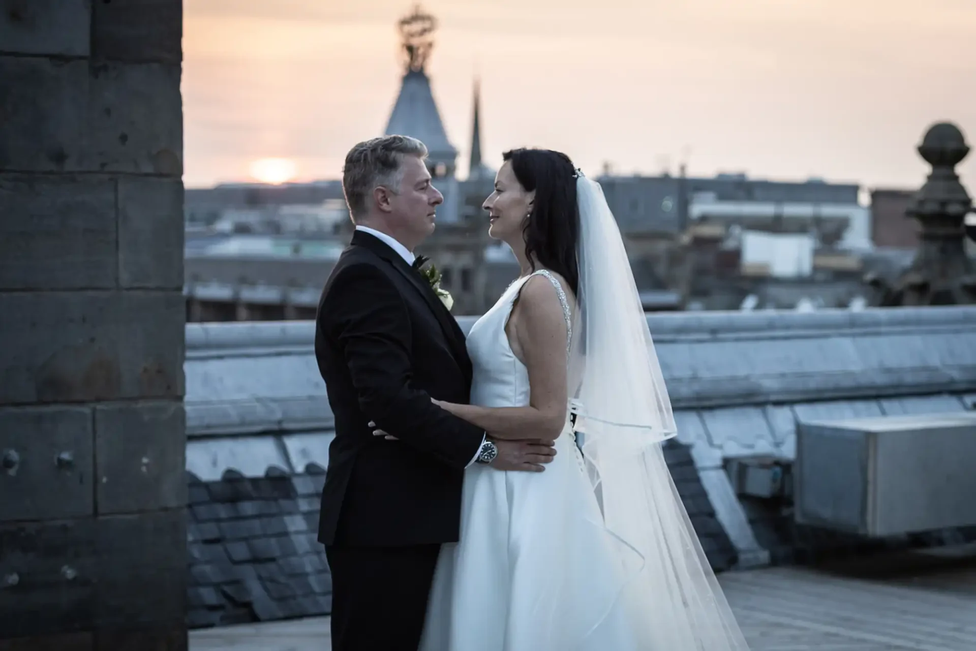 A bride and groom embrace on a rooftop at sunset, with city buildings in the background.