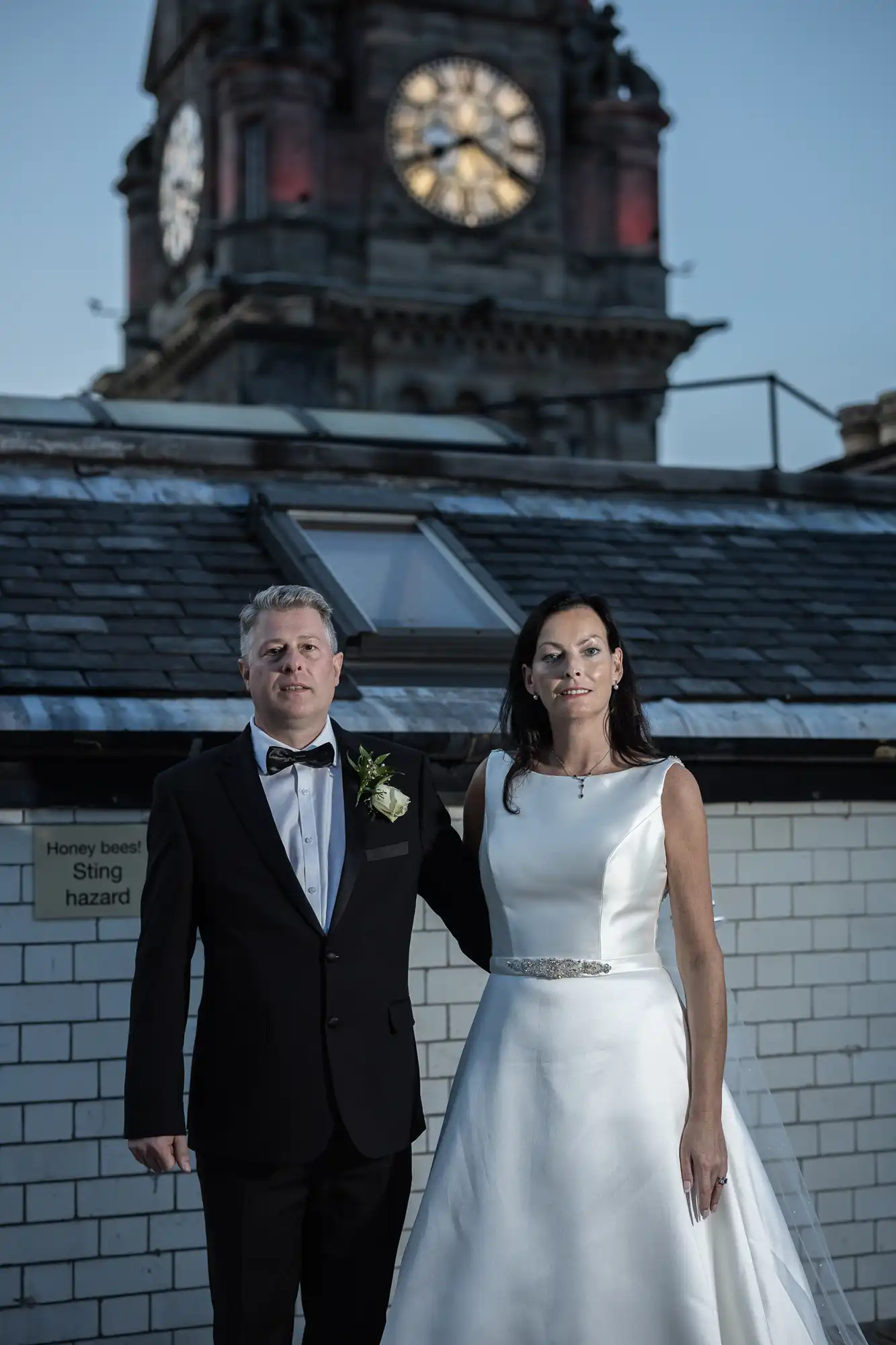 A bride and groom stand in formal wedding attire before a large clock tower under a clear sky.