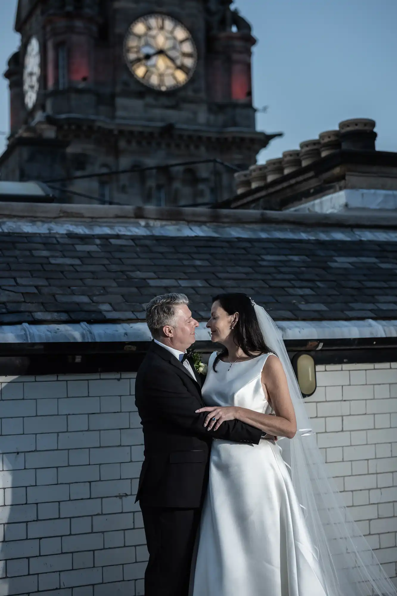 A bride and groom share a moment, standing close together with a historical clock tower in the soft-lit background.