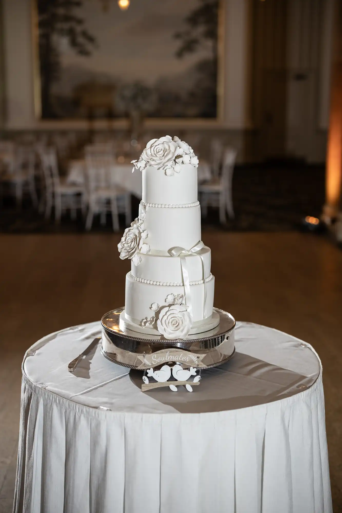 A four-tier white wedding cake with floral and ribbon decorations, displayed on a silver stand on a draped table in an elegant room.