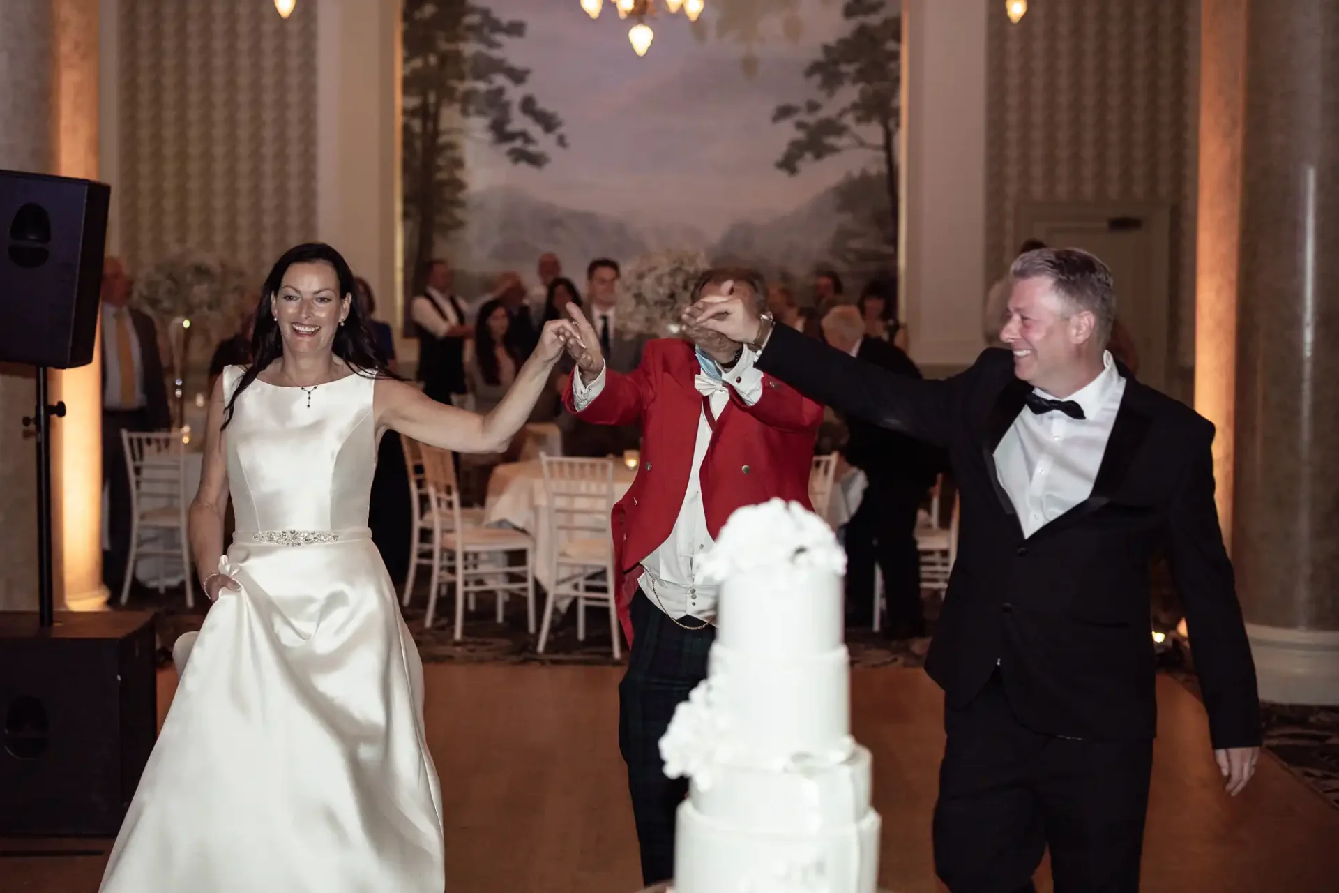 A joyful bride and groom dancing at their wedding reception with a guest wearing a red jacket, near a white wedding cake.