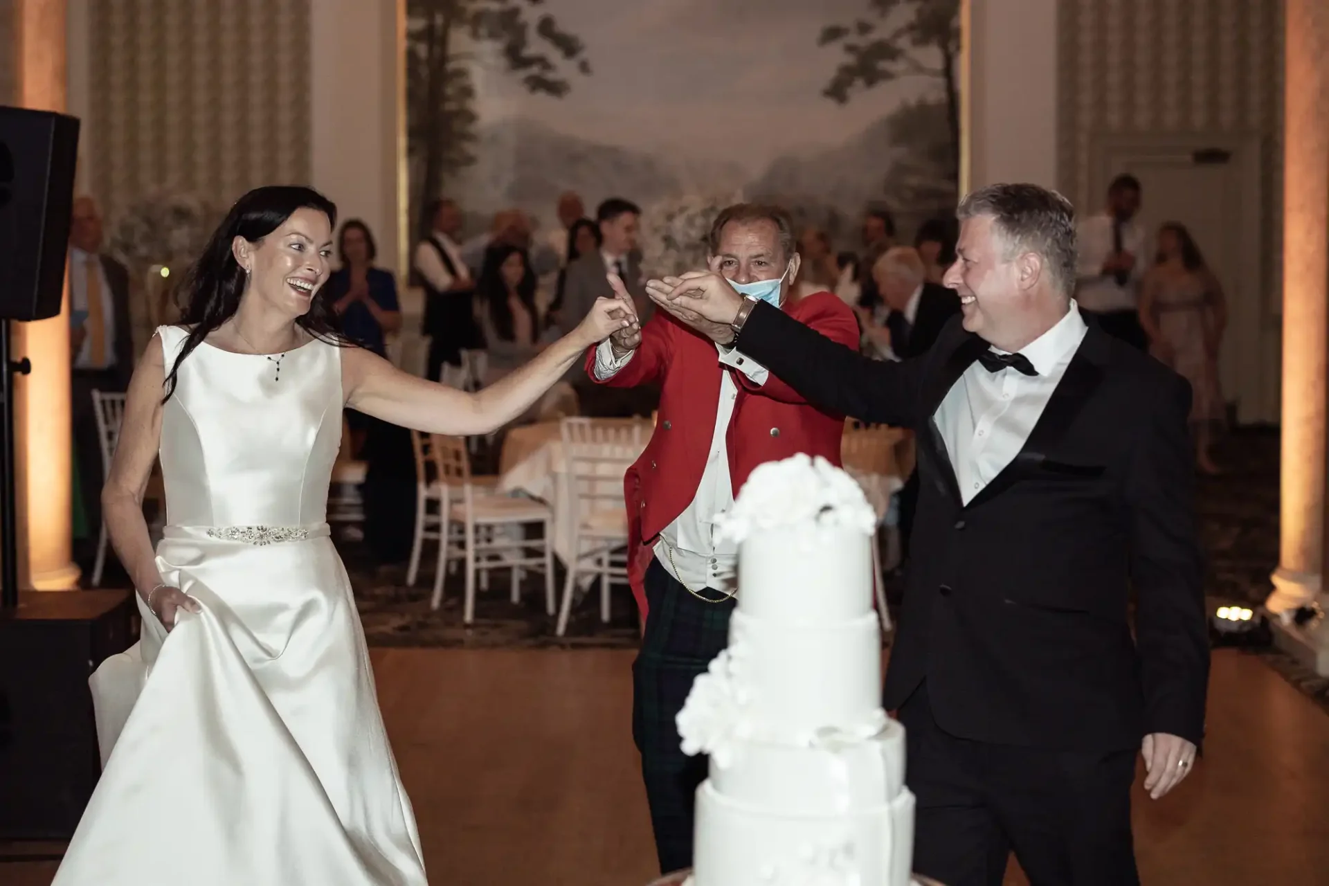 A bride in a white gown fist-bumping an older man in a red jacket, while a man in a black tuxedo smiles beside them at a wedding reception.