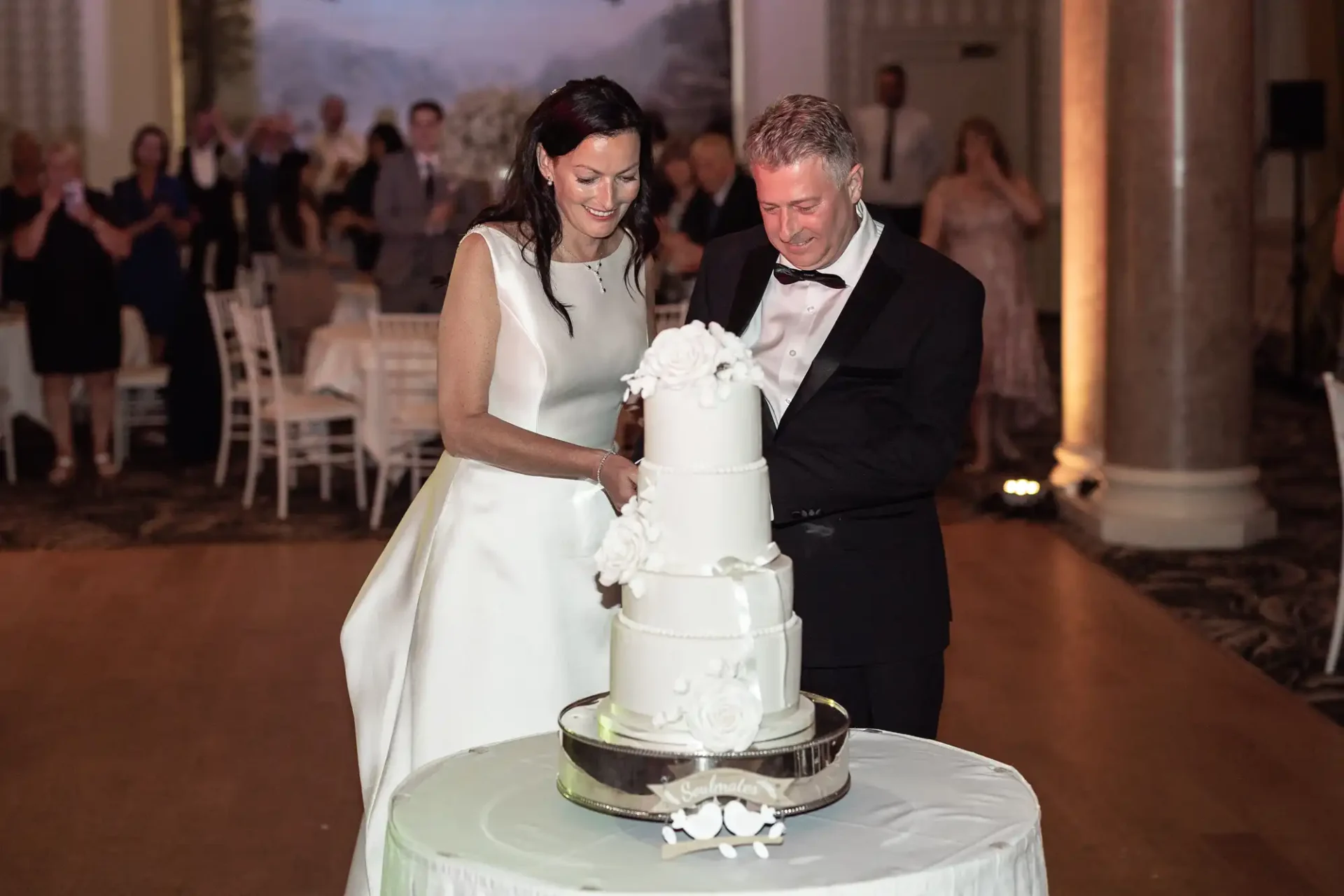 A couple in formal attire smiling as they cut a three-tiered white wedding cake at a reception.
