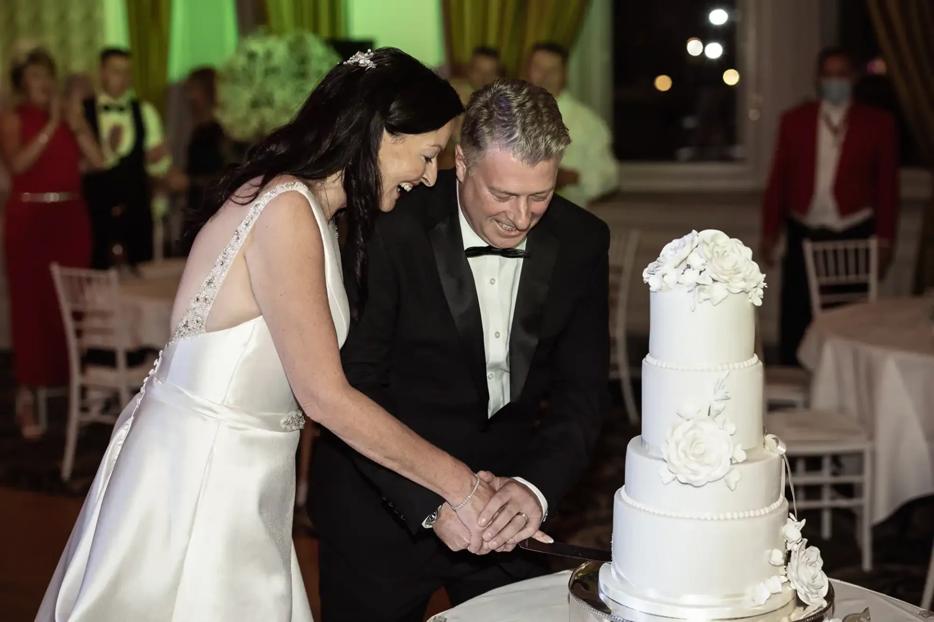 A bride and groom smiling as they cut a multi-tiered white wedding cake together at their reception.