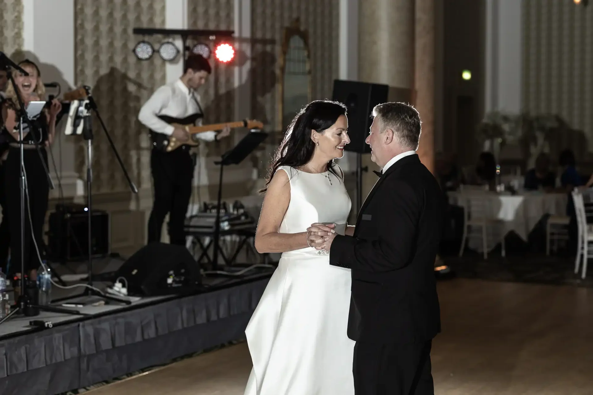 A couple dancing closely in a formal setting during a wedding reception, with a live band performing in the background.
