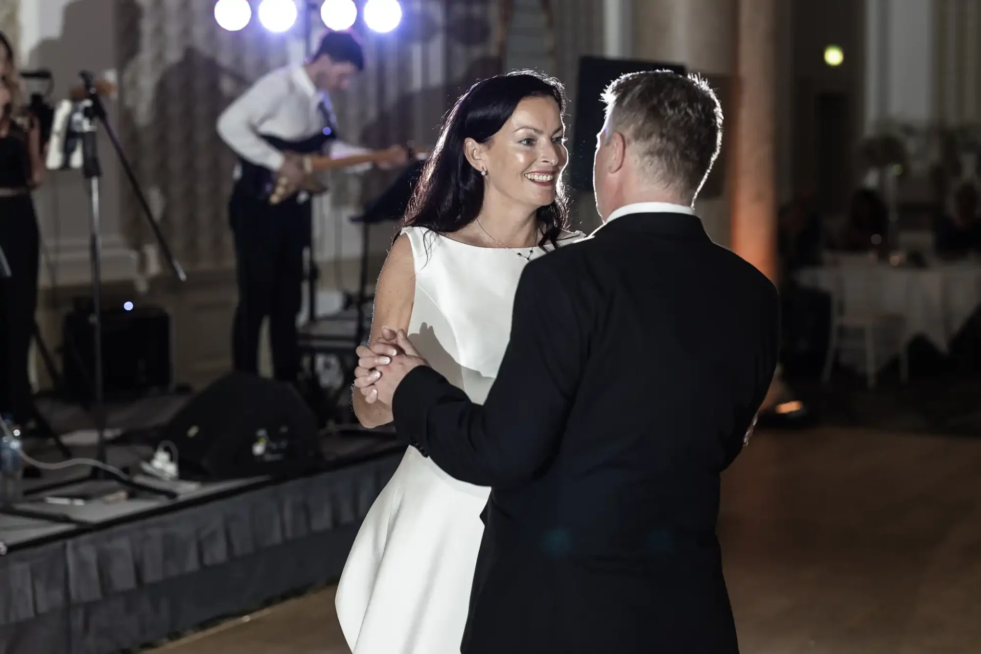 A couple in formal attire smiling at each other while dancing at an event, with a band performing in the background.