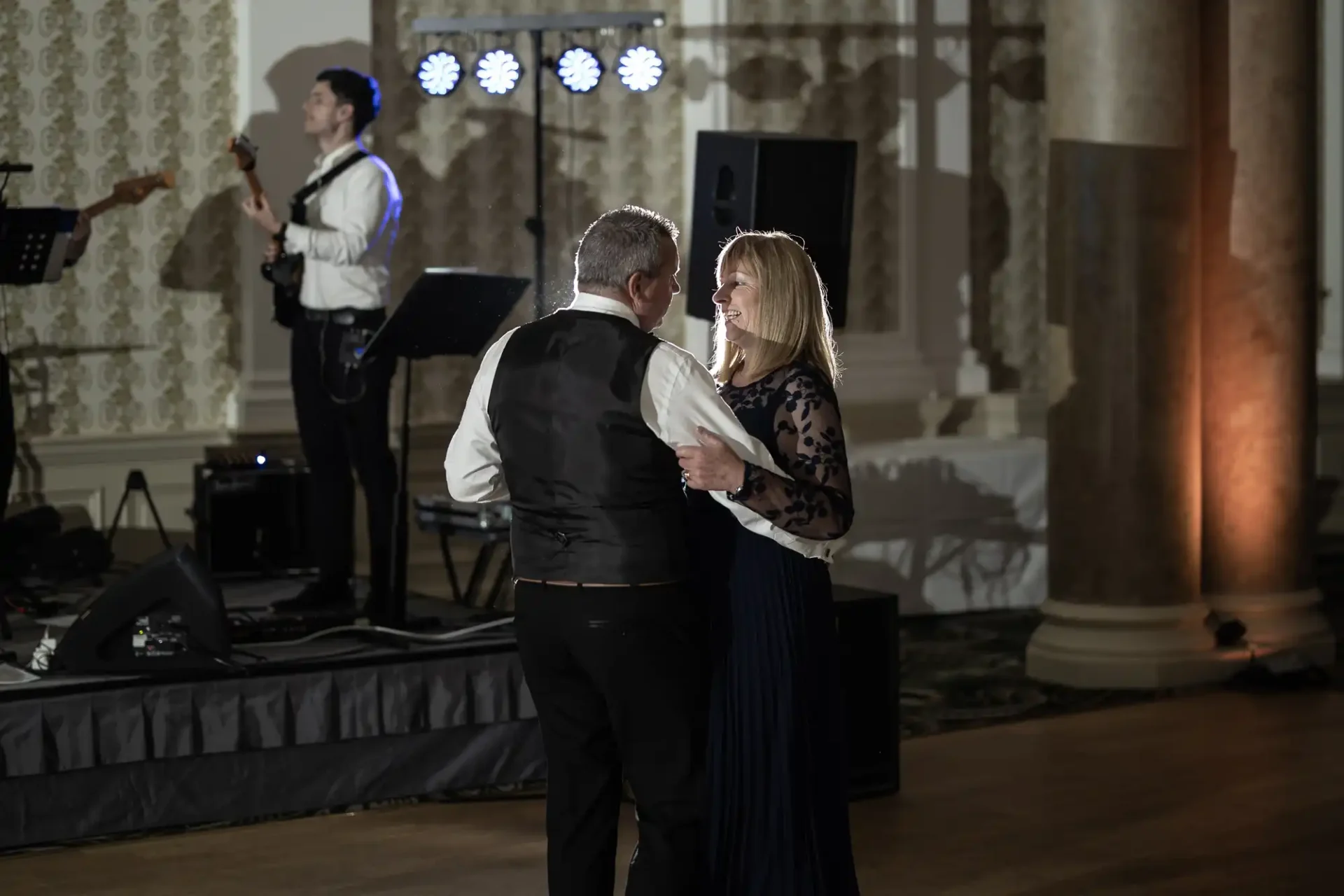 A couple dances closely at an elegant event while a musician performs in the background under blue lights.