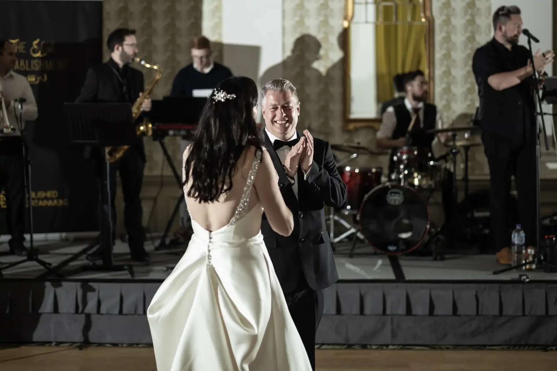 A bride and groom share a dance, smiling joyfully, with a live band performing in the background in an elegant ballroom.