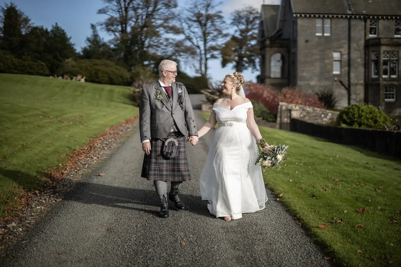 A bride in a white dress and a groom in a grey jacket and tartan kilt walk hand in hand on a path beside a large stone building and green lawn.