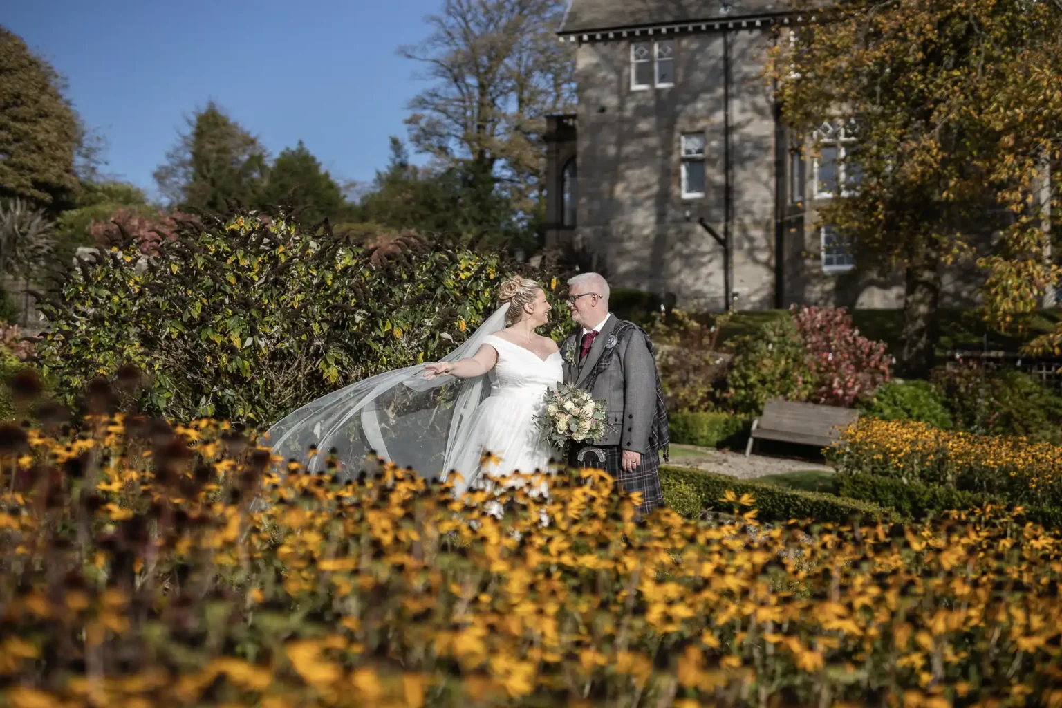A bride and groom stand together in a garden filled with yellow flowers, with a historic building and trees in the background on a sunny day.