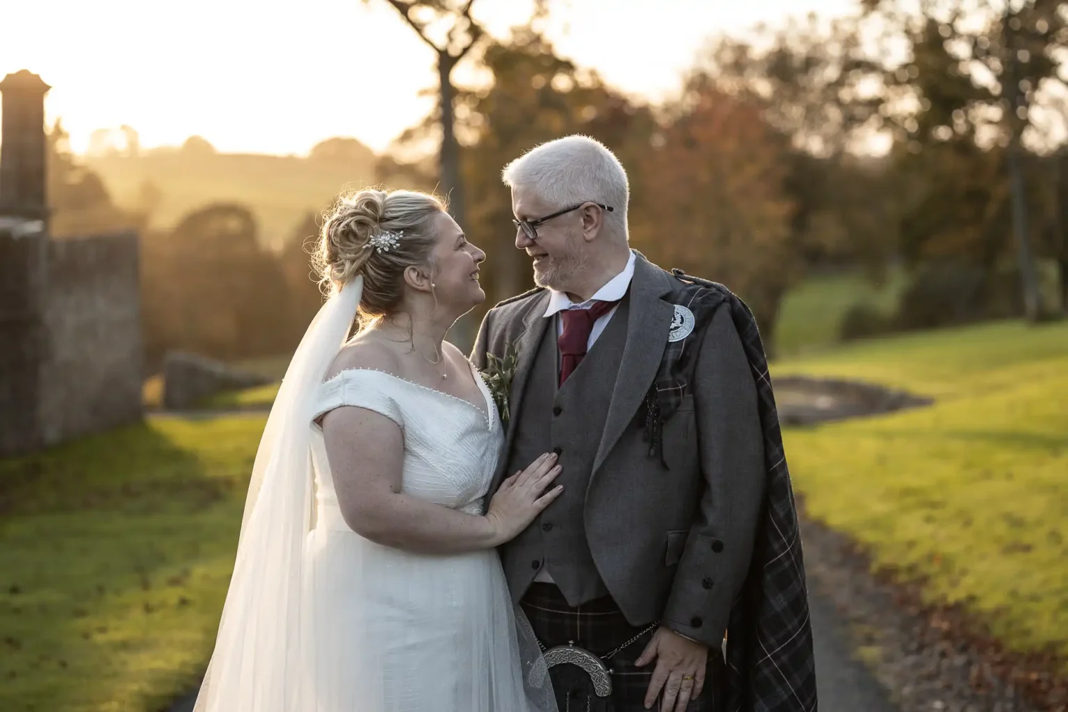 A bride in a white dress and a groom in a grey suit with a plaid sash stand outdoors, looking at each other and smiling, with green grass and trees in the background.