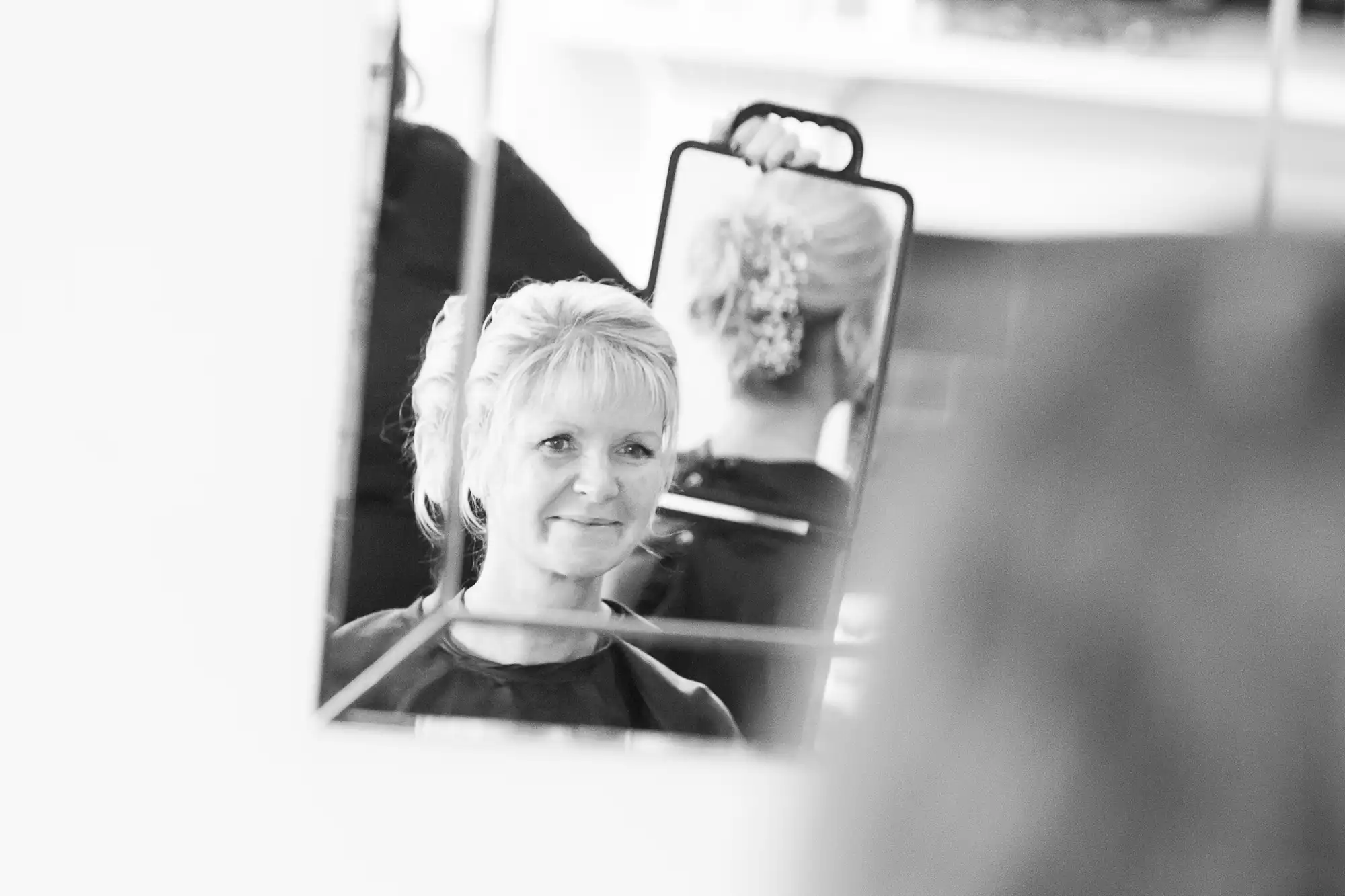 A middle-aged woman smiling subtly at her reflection in a handheld mirror, captured in black and white.