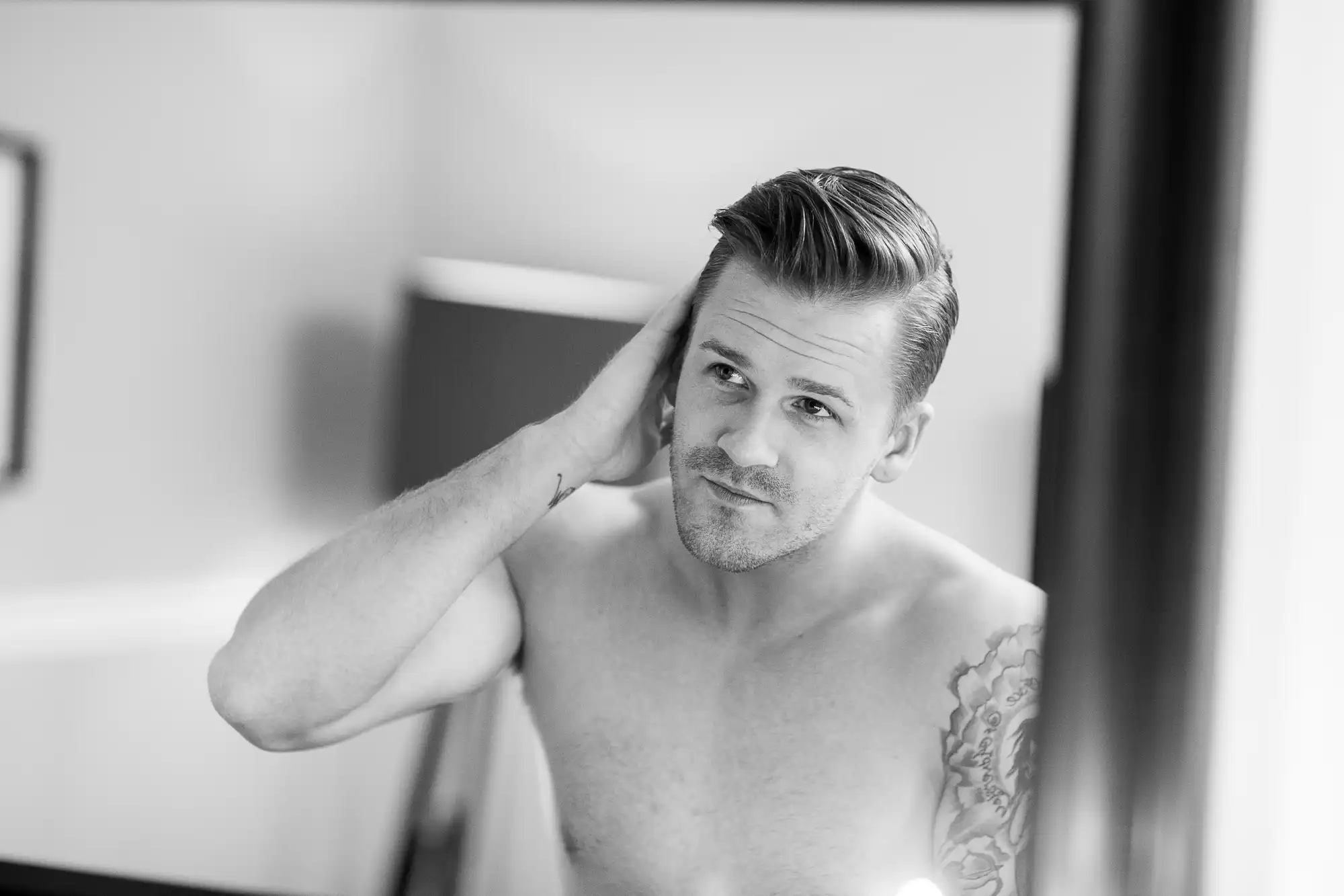 Black-and-white photo of a shirtless man with a slicked-back hairstyle and a tattoo, looking at his reflection in a mirror and touching his hair.