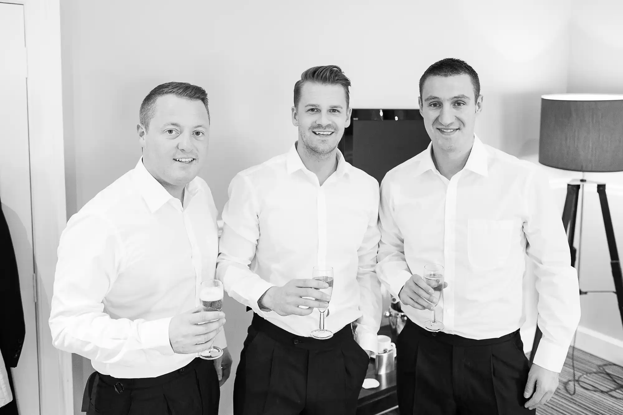 Three men in white shirts holding champagne glasses, smiling in a black and white photo in an indoor setting.
