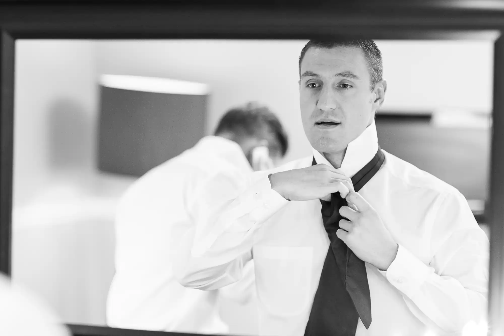 Man in a white shirt adjusts his black tie while looking at his reflection in a mirror, depicted in a black and white photograph.