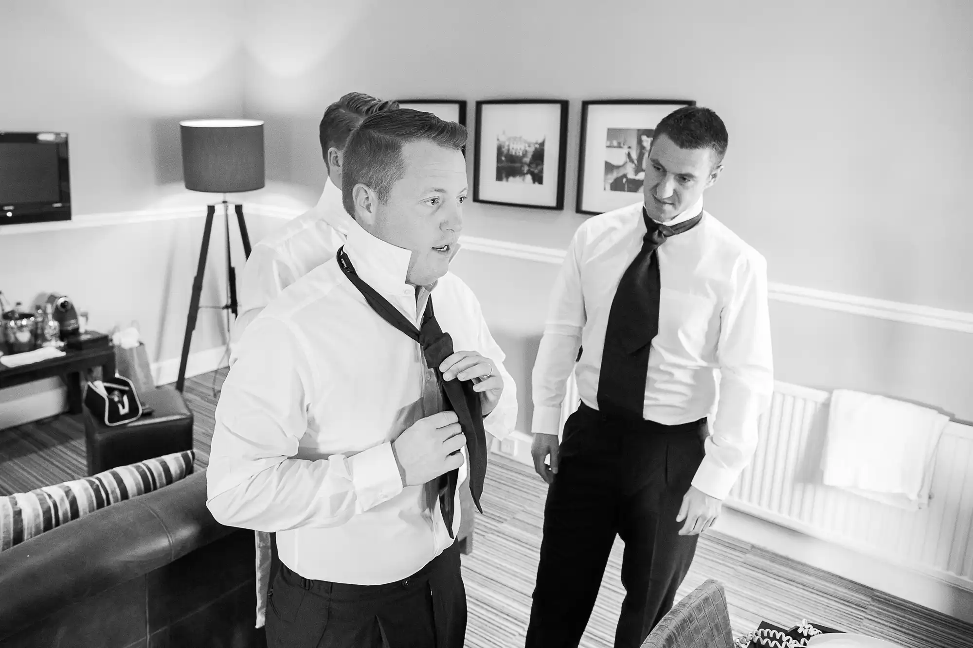 Two men in a well-lit room dressing up in formal attire with one adjusting his tie while the other watches. room decor includes photographs on the wall and a standing lamp.