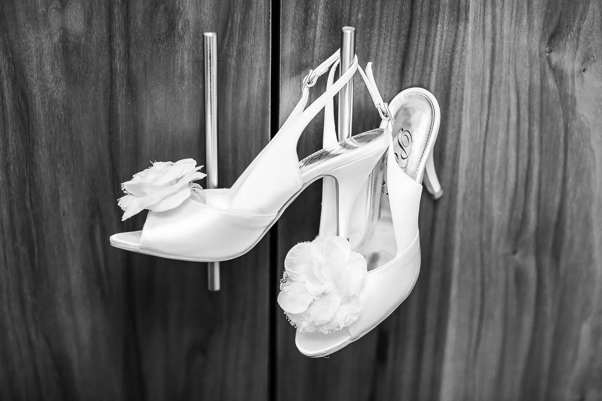 A pair of white bridal high heel shoes with floral decorations hanging on a wooden fence.