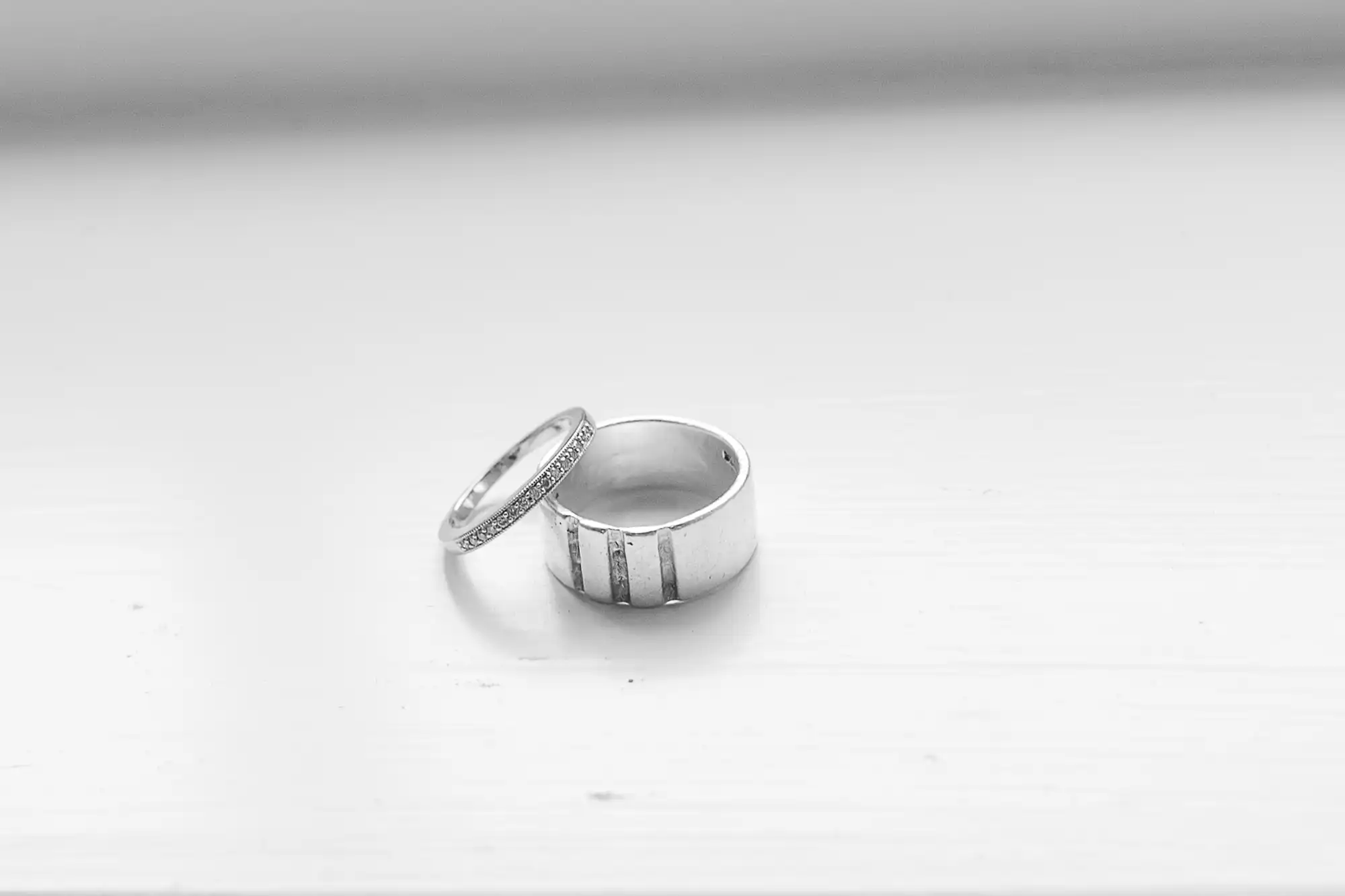Two rings on a plain surface, one slender with small embedded stones and the other wider with groove details, presented in a monochrome setting.