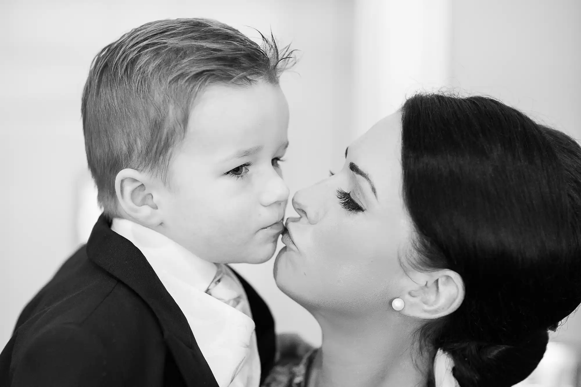 A black and white photo of a young boy in a suit receiving a kiss on the cheek from a woman with long eyelashes.