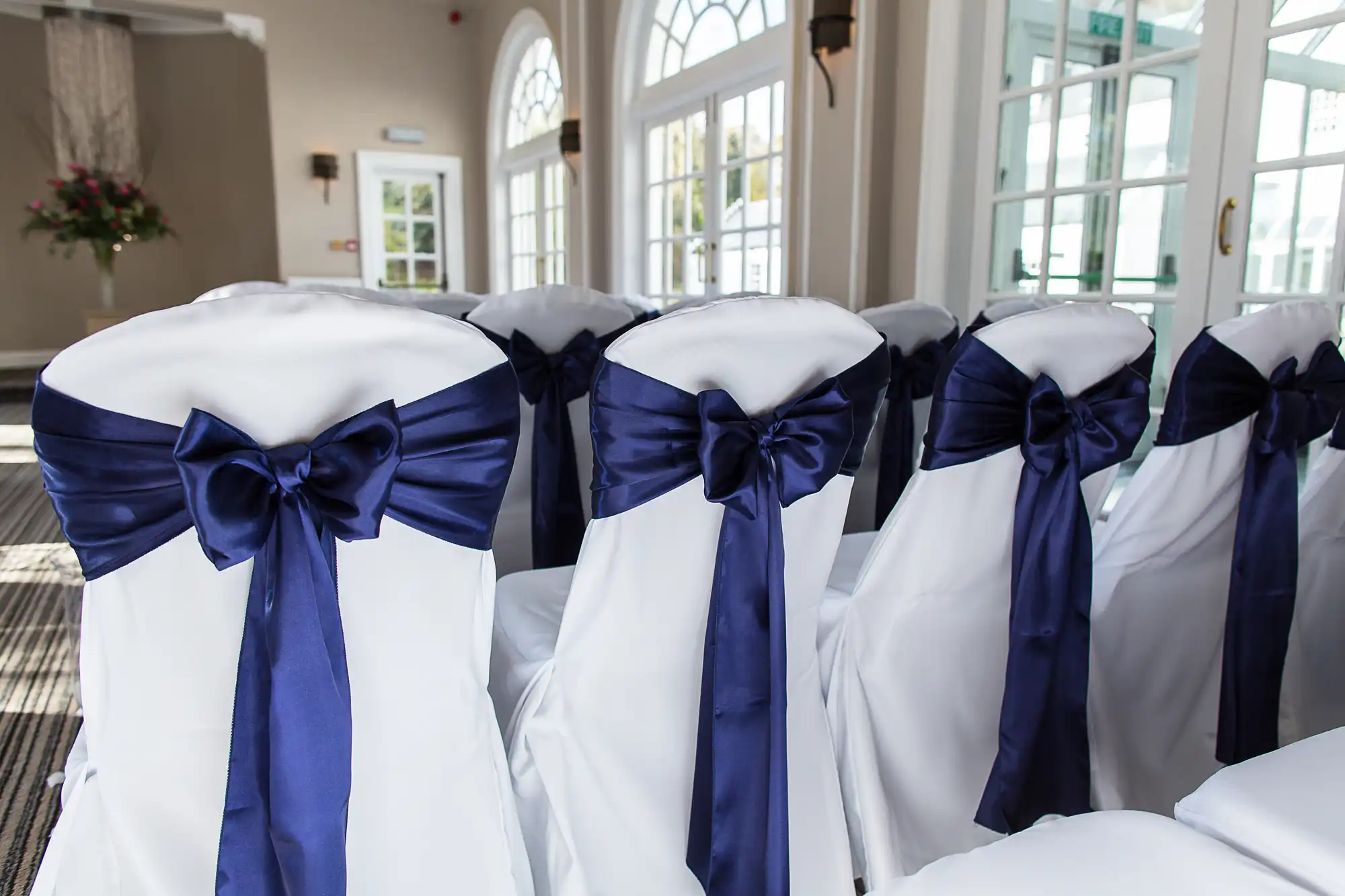White chairs with dark blue satin bows lined up in a bright room with windows and a floral arrangement in the background.